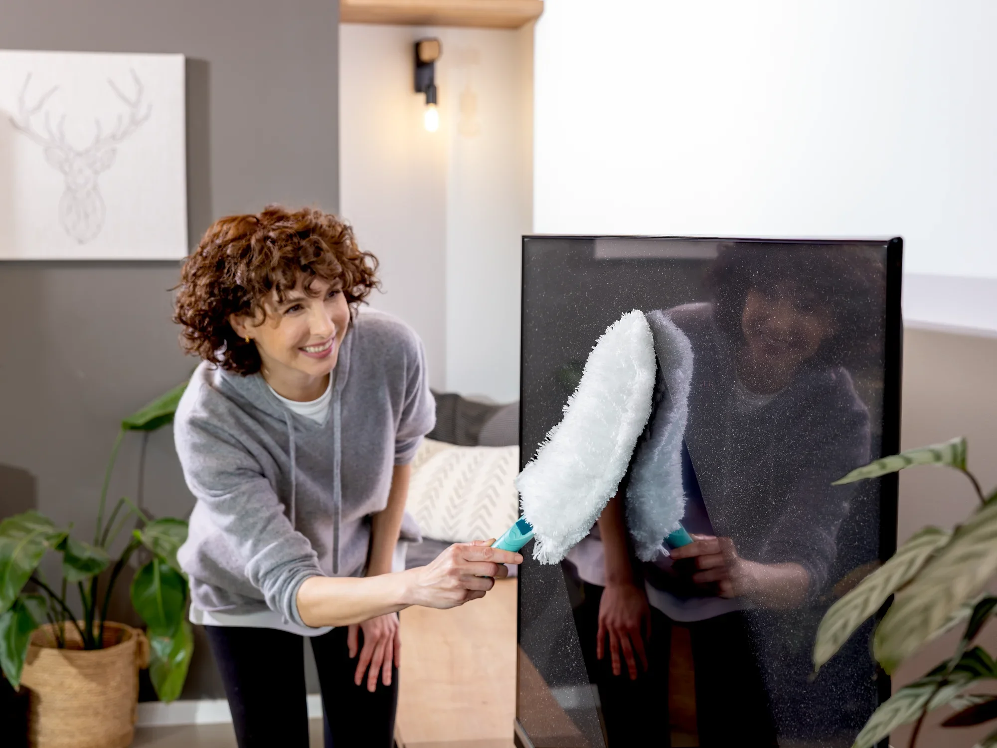 Een persoon gebruikt de SUPERDUSTER XL plumeau om een groot scherm schoon te maken. De plumeau heeft een witte, pluizige kop en een blauw handvat.