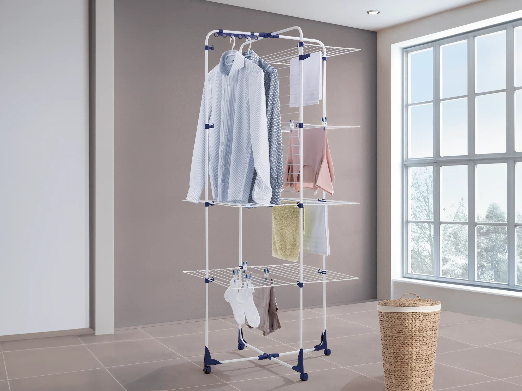 Séchoir à tour Classic Tower 450 : un séchoir à linge blanc à plusieurs niveaux avec des accents bleus, chargé de vêtements, placé dans une pièce lumineuse à côté d'une fenêtre et d'un panier à linge.