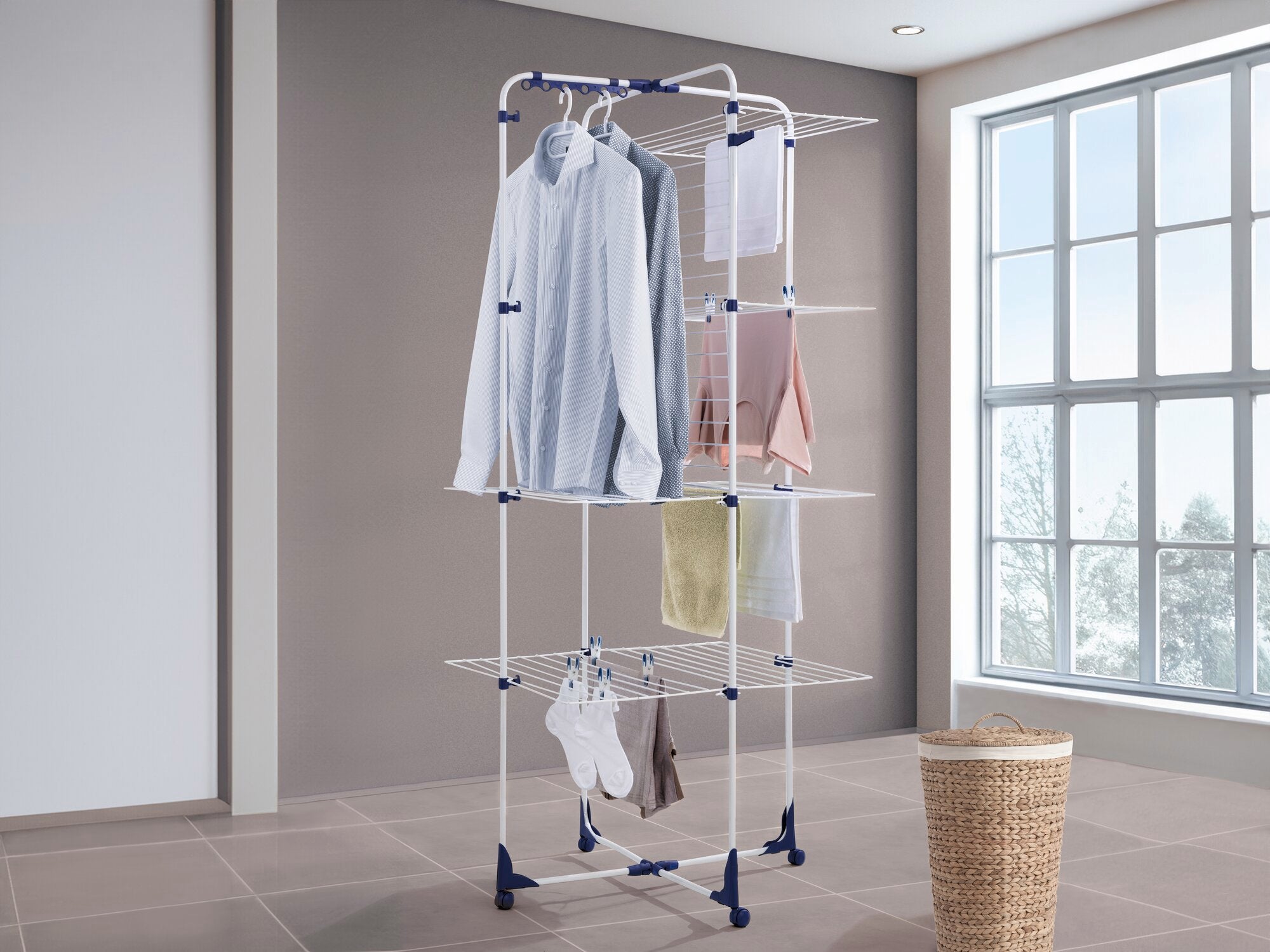 Classic Tower 450 torendroger: Een wit droogrek met meerdere niveaus en blauwe accenten, vol met kleding, staat in een lichte kamer naast een raam en een wasmand.