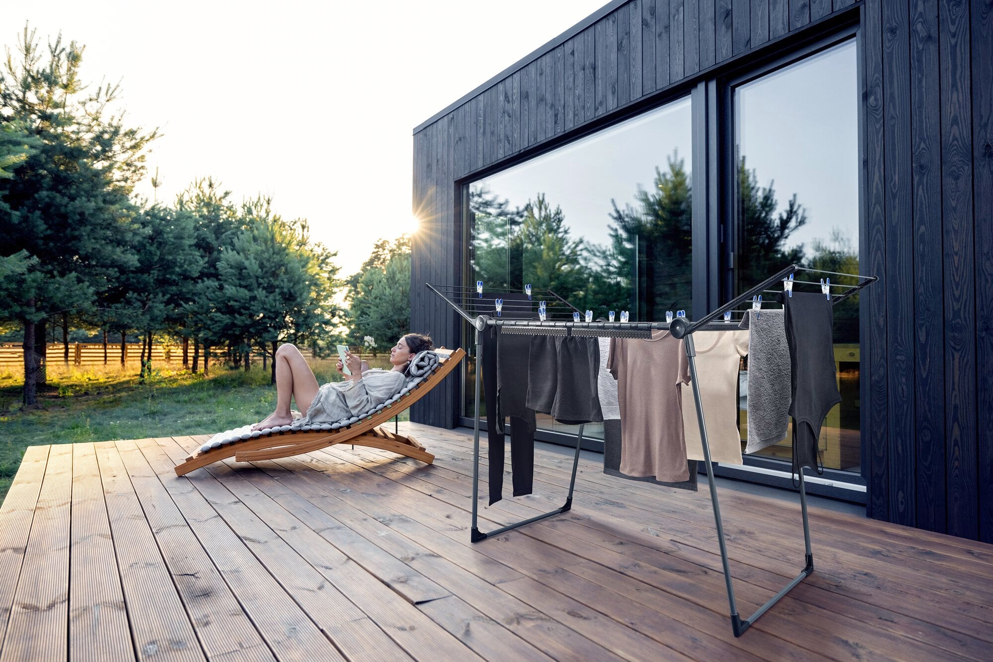 Una moderna secadora de pie Pegasus 180 Solid se encuentra en una terraza de madera frente a una elegante casa. Al fondo, una persona se relaja en una tumbona.