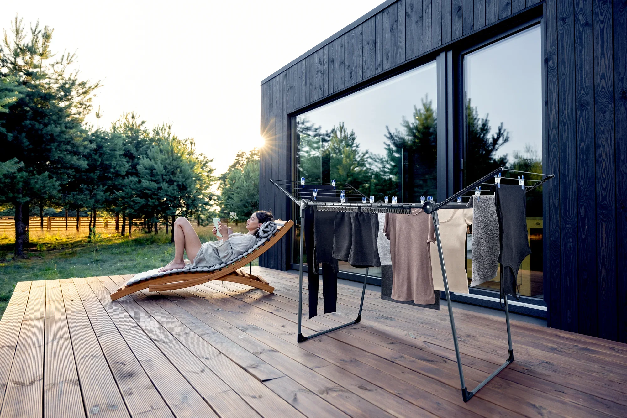 A modern Pegasus 180 Solid freestanding dryer stands on a wooden terrace in front of a stylish house. In the background, a person relaxes on a lounger.