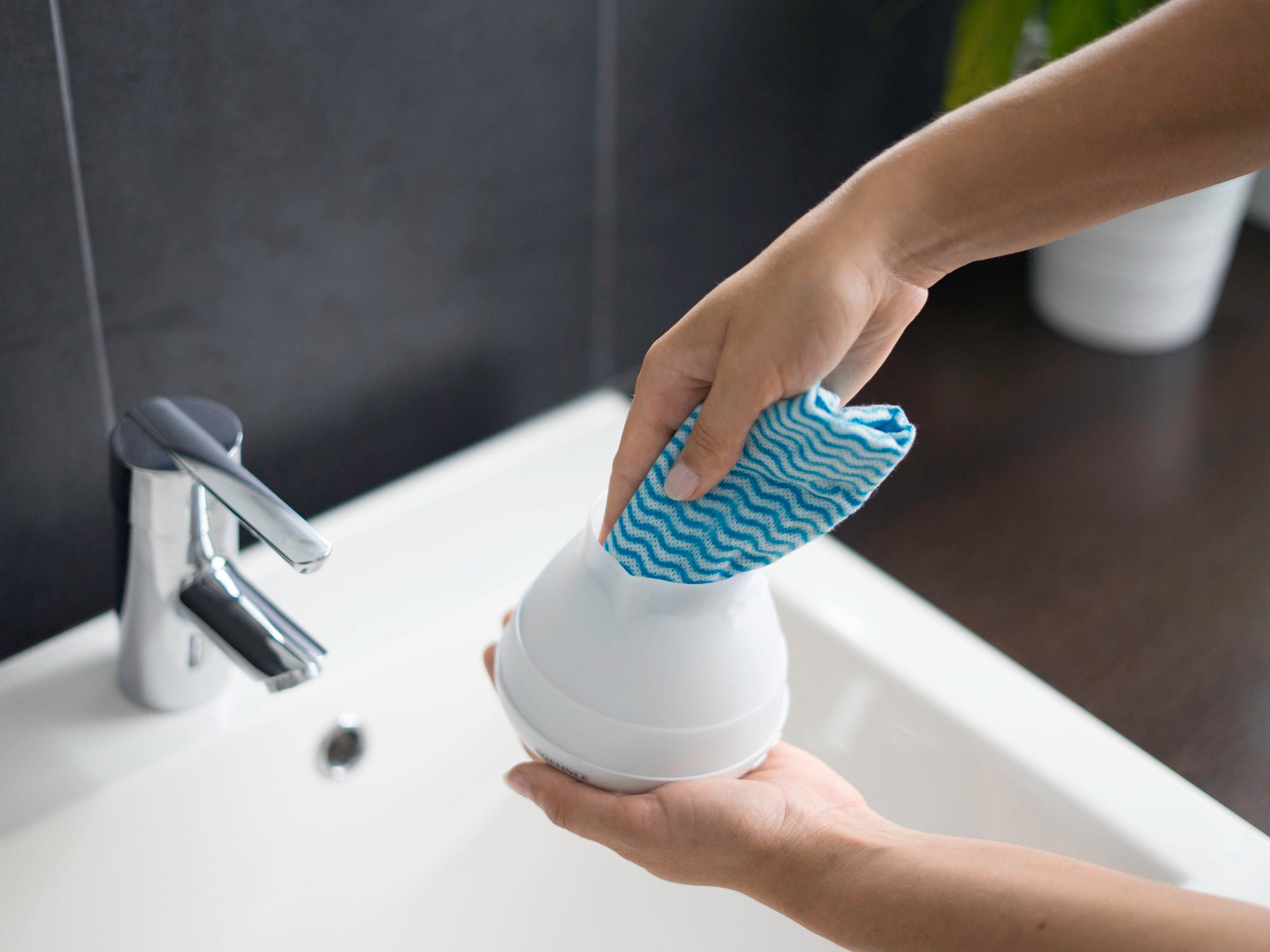 Eine Person reinigt den Design Aroma Diffuser Bari mit einem blauen Tuch über einem Waschbecken. Der Diffuser hat eine glatte, weiße Oberfläche und wird sorgfältig abgewischt.