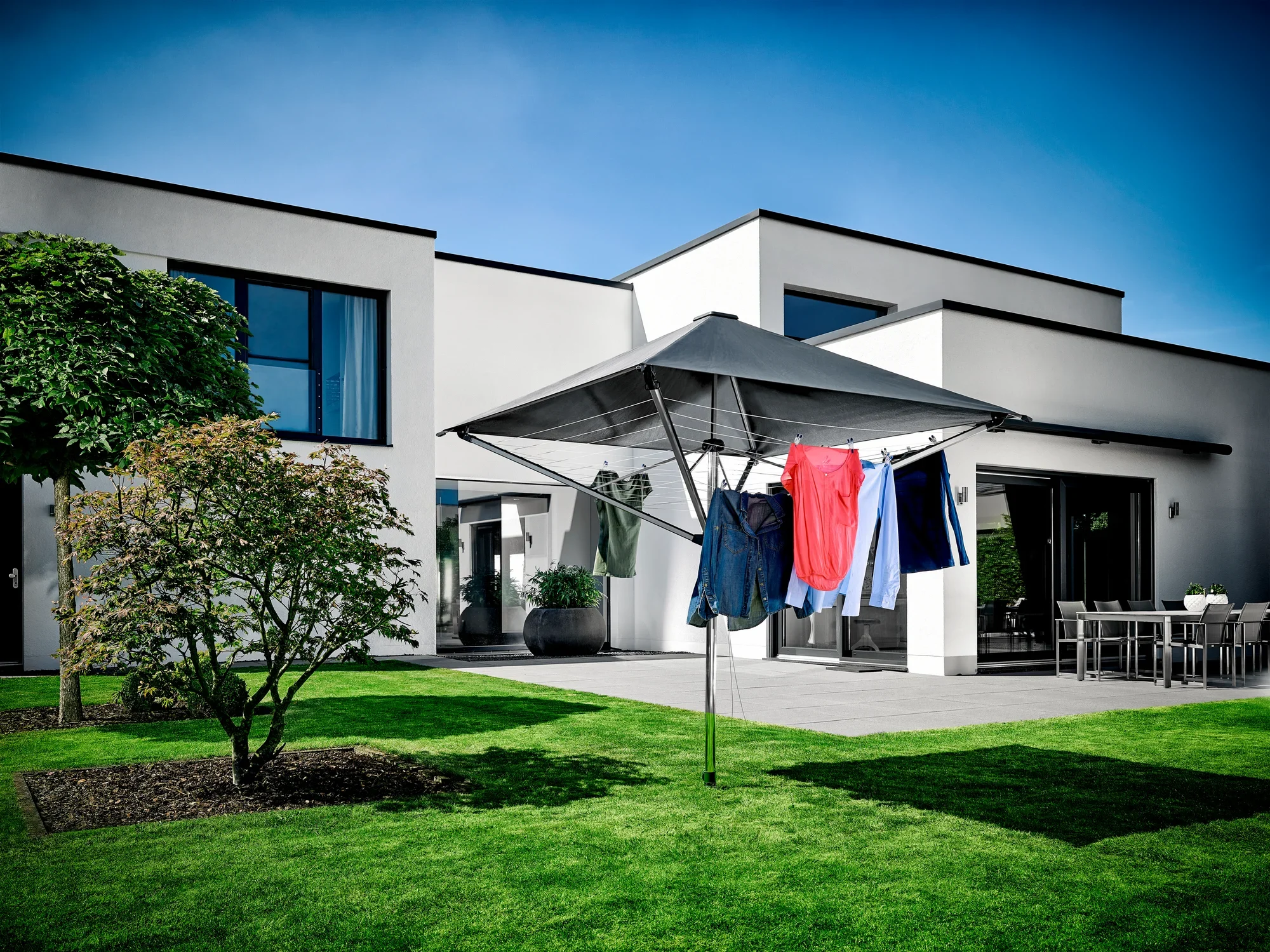 Séchoir à linge LinoProtect 400 dans le jardin devant une maison moderne. Le séchoir à linge est ouvert et recouvert de vêtements colorés. Le ciel est clair et bleu.