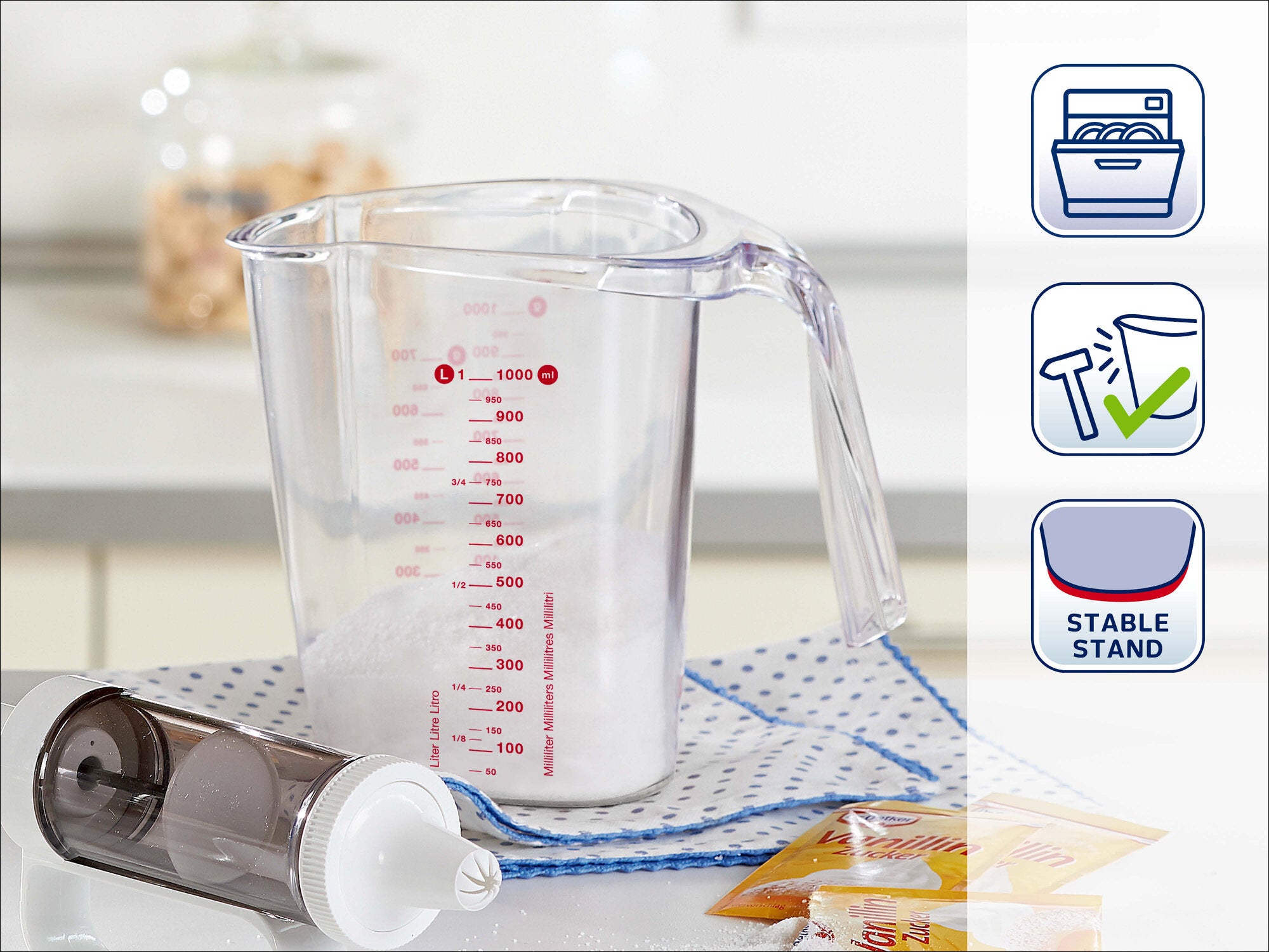 Leifheit Messbecher mit roter Milliliter-Skala und Ausgießer auf einer Küchenarbeitsplatte. Daneben liegen Spritztüllen und Zubehör; Symbole zeigen Spülmaschineneignung und sicheren Stand.