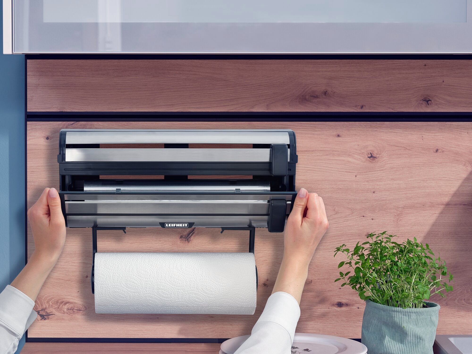 Parat Royal wandrolhouder van Leifheit gemonteerd op een houten wand. Twee handen bedienen de houder terwijl er een keukenrol onder hangt. Ernaast staat een pot met kruiden.