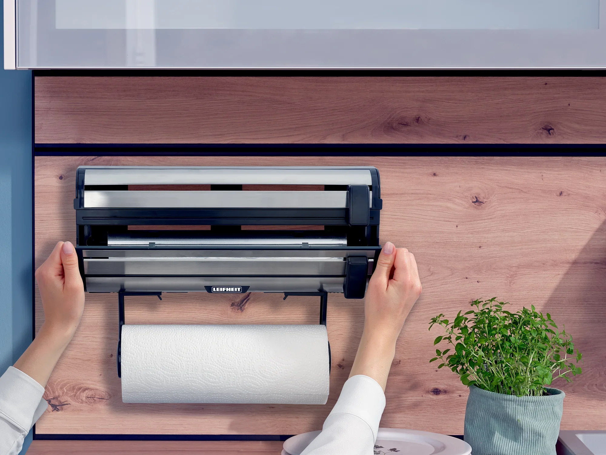 Uchwyt ścienny na rolki Parat Royal firmy Leifheit zamontowany na drewnianej ścianie. Dwie ręce obsługują uchwyt, podczas gdy rolka kuchenna wisi poniżej. Obok znajduje się doniczka z ziołami.