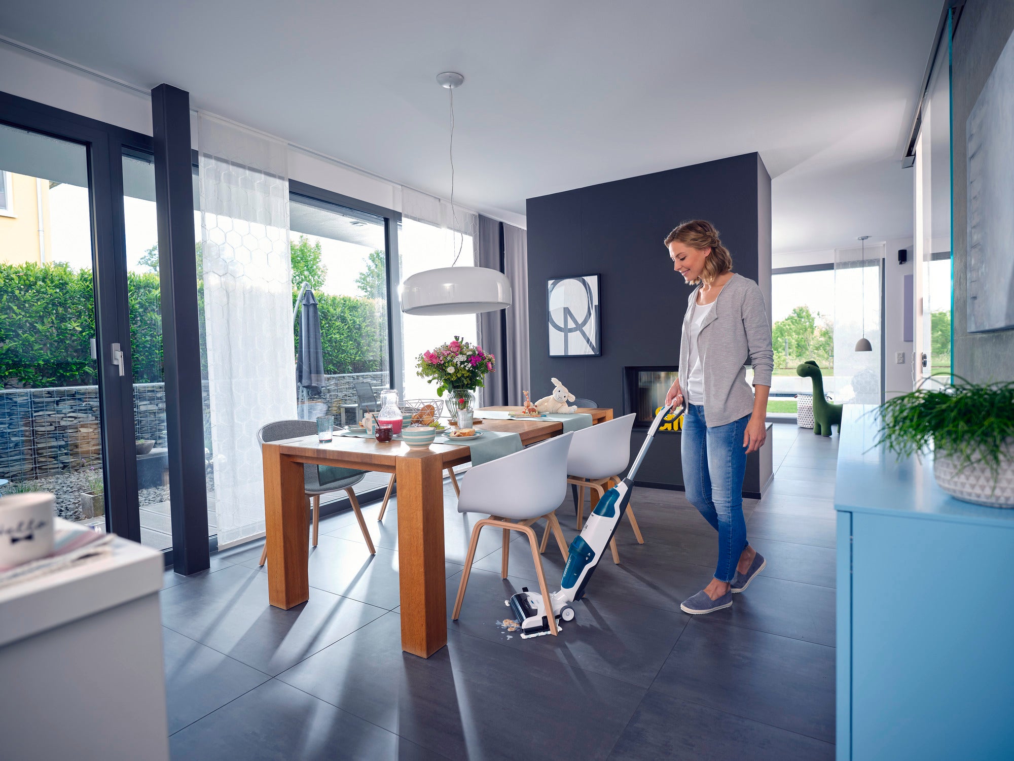 Une femme utilise l'aspirateur Regulus Aqua PowerVac Pro dans une salle à manger moderne et lumineuse, avec de grandes fenêtres et une table en bois élégante.