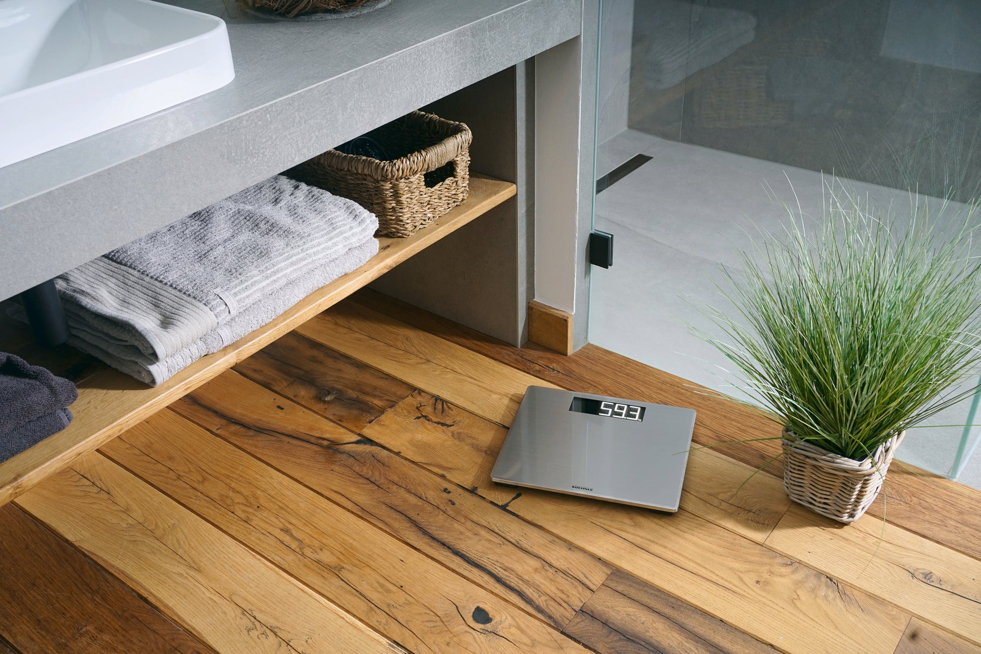 A Style Sense Safe 300 digital bathroom scale lies on an elegant wooden floor in the bathroom. Next to it is a basket with towels and a plant.