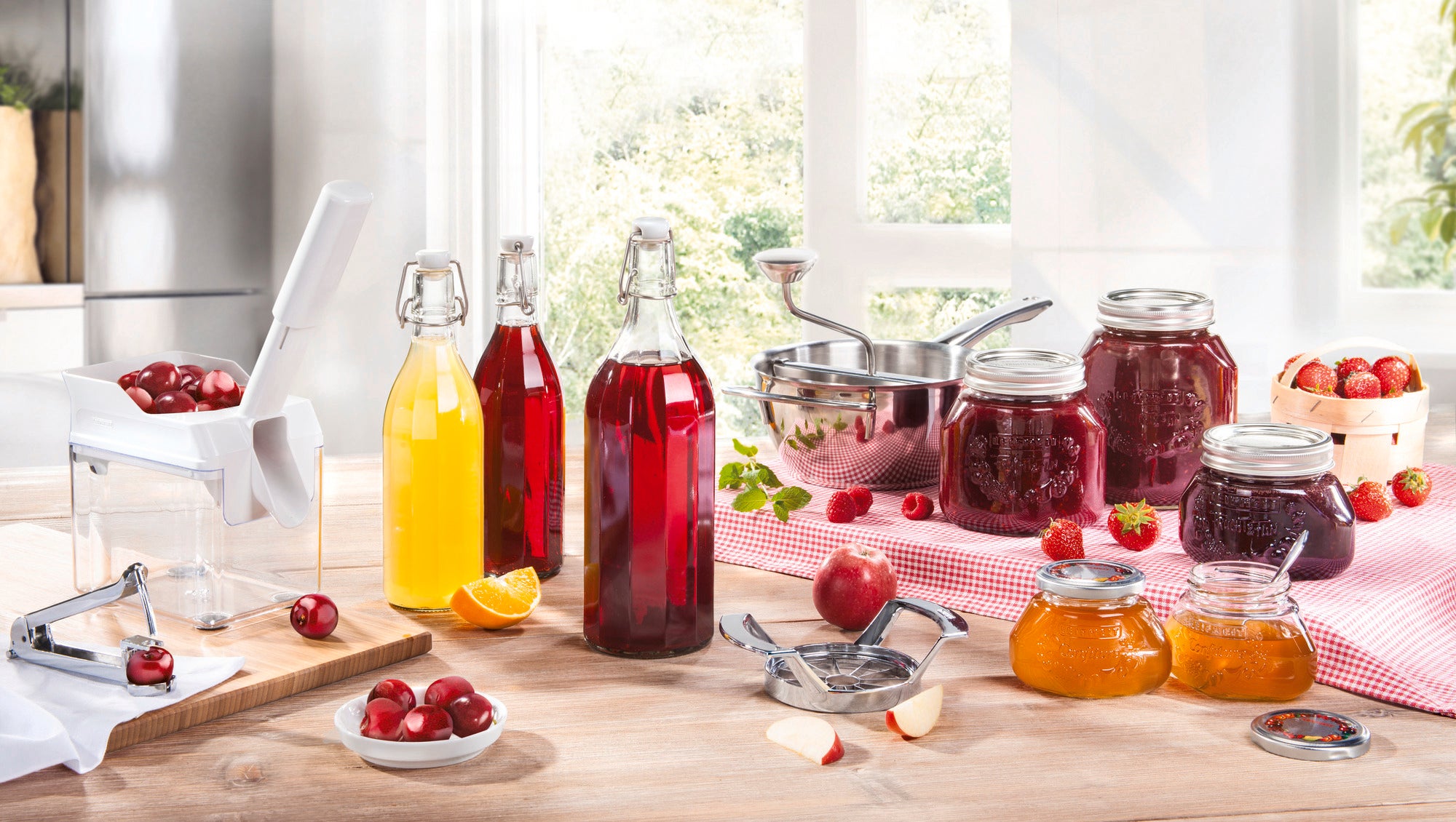 A Leifheit cherry pitter stands on a table surrounded by jars of cherry jam, juice bottles and fresh cherries. A bright kitchen can be seen in the background.