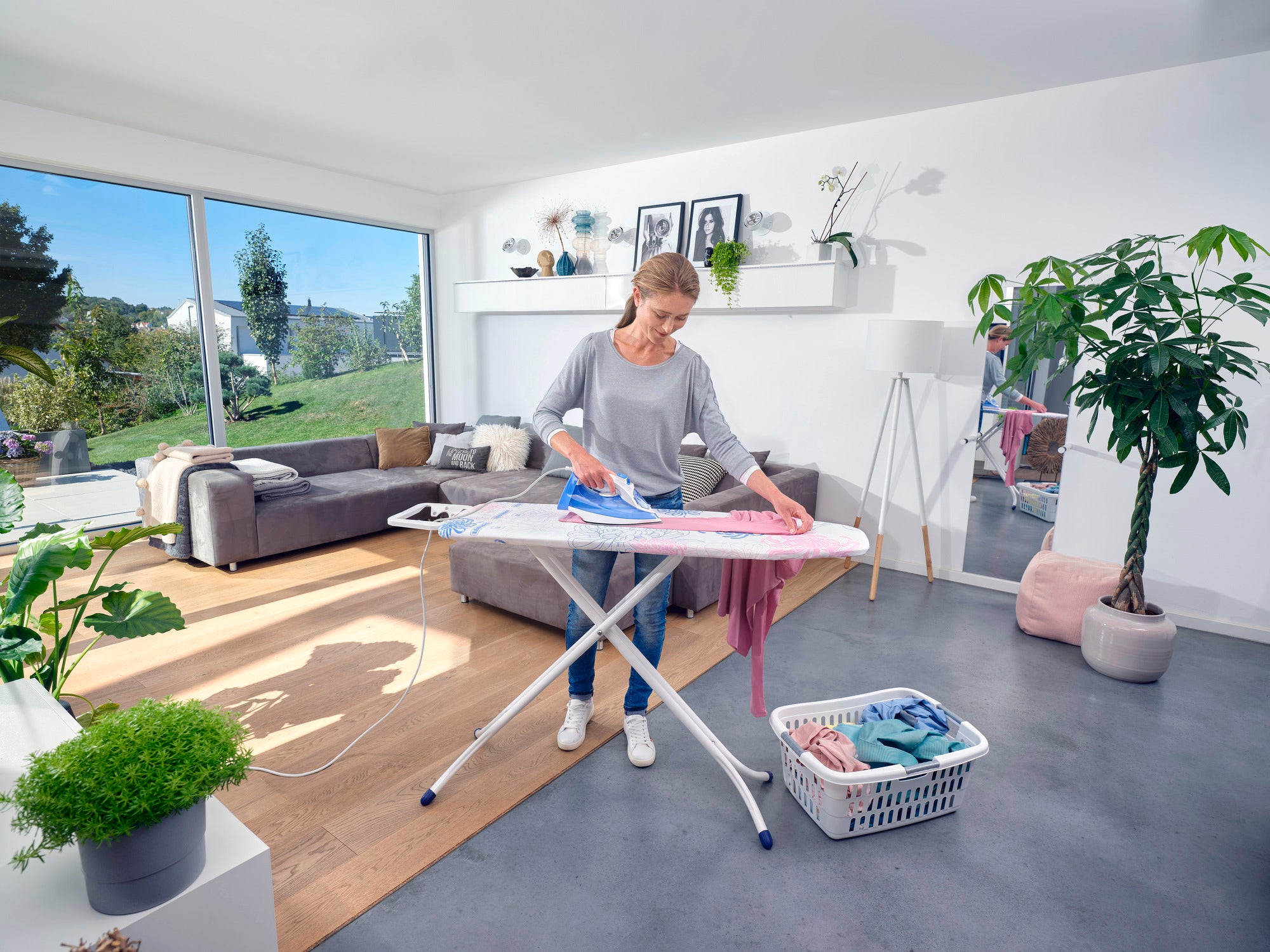 Eine Frau bügelt Kleidung auf einem Bügeltisch Classic M Compact in einem modernen Wohnzimmer. Der Raum ist hell und stilvoll eingerichtet mit Pflanzen und großen Fenstern.
