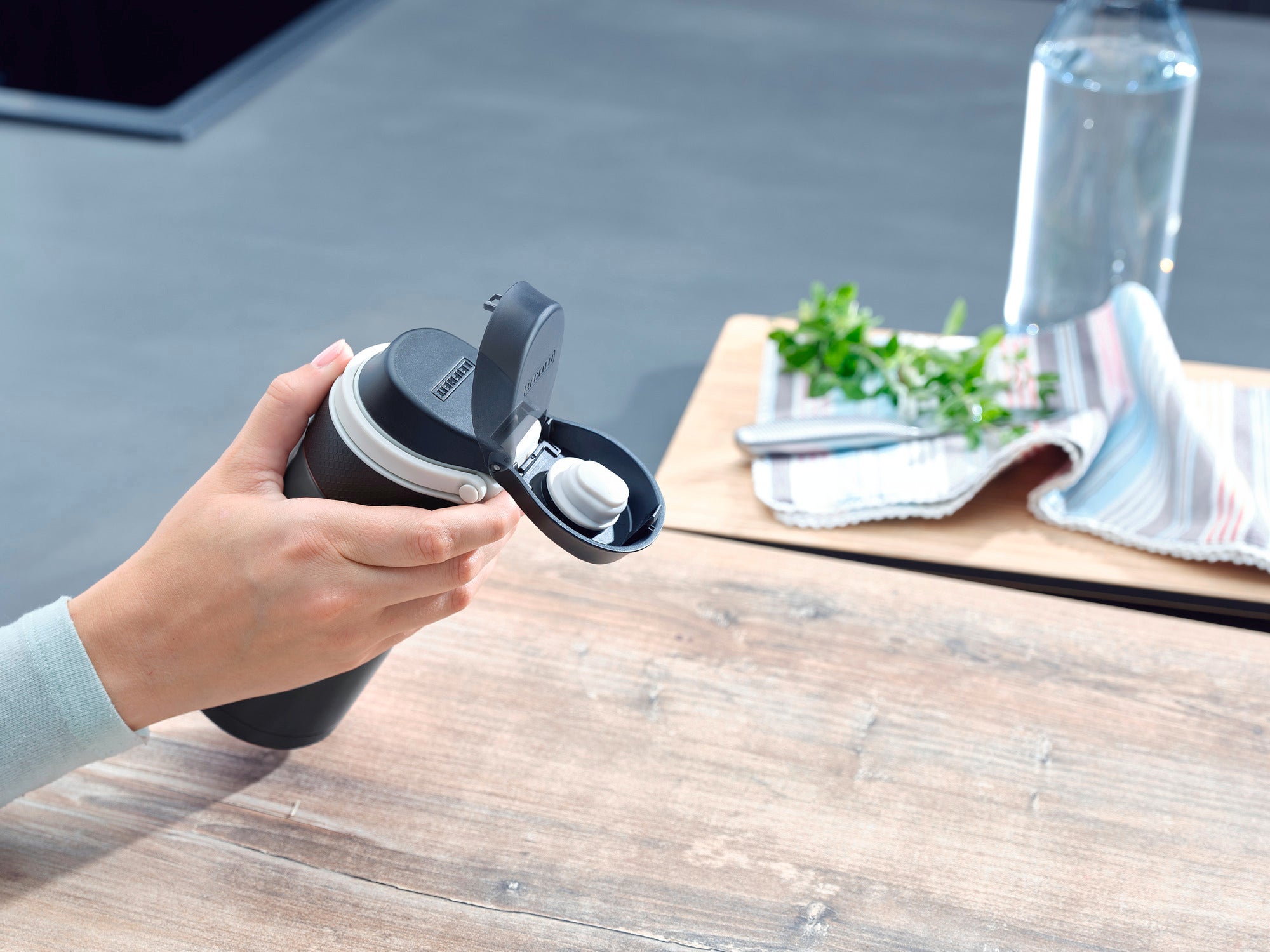 Une main tient un mug isotherme Flip noir d'une contenance de 350 ml. Le mug est doté d'un couvercle rabattable pratique. À l'arrière-plan, on aperçoit une table, une bouteille d'eau et des herbes aromatiques.