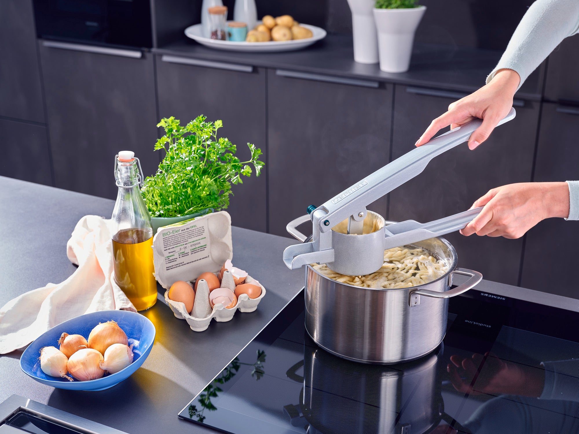 Eine Person verwendet eine Kartoffel-Spätzlepresse, um frische Spätzle in einen Topf zu pressen. Auf der Arbeitsfläche stehen Eier, Öl und frische Kräuter bereit.