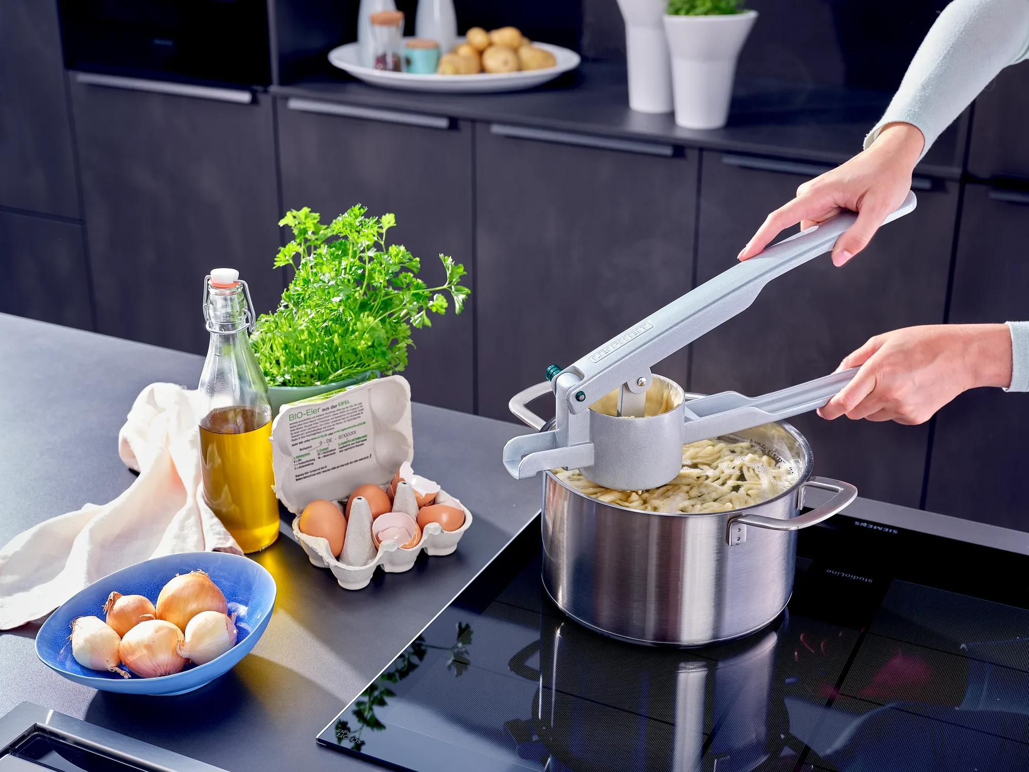 Osoba używa prasy do ziemniaków spaetzle, aby wycisnąć świeże spaetzle do garnka. Jajka, olej i świeże zioła są gotowe na powierzchni roboczej.