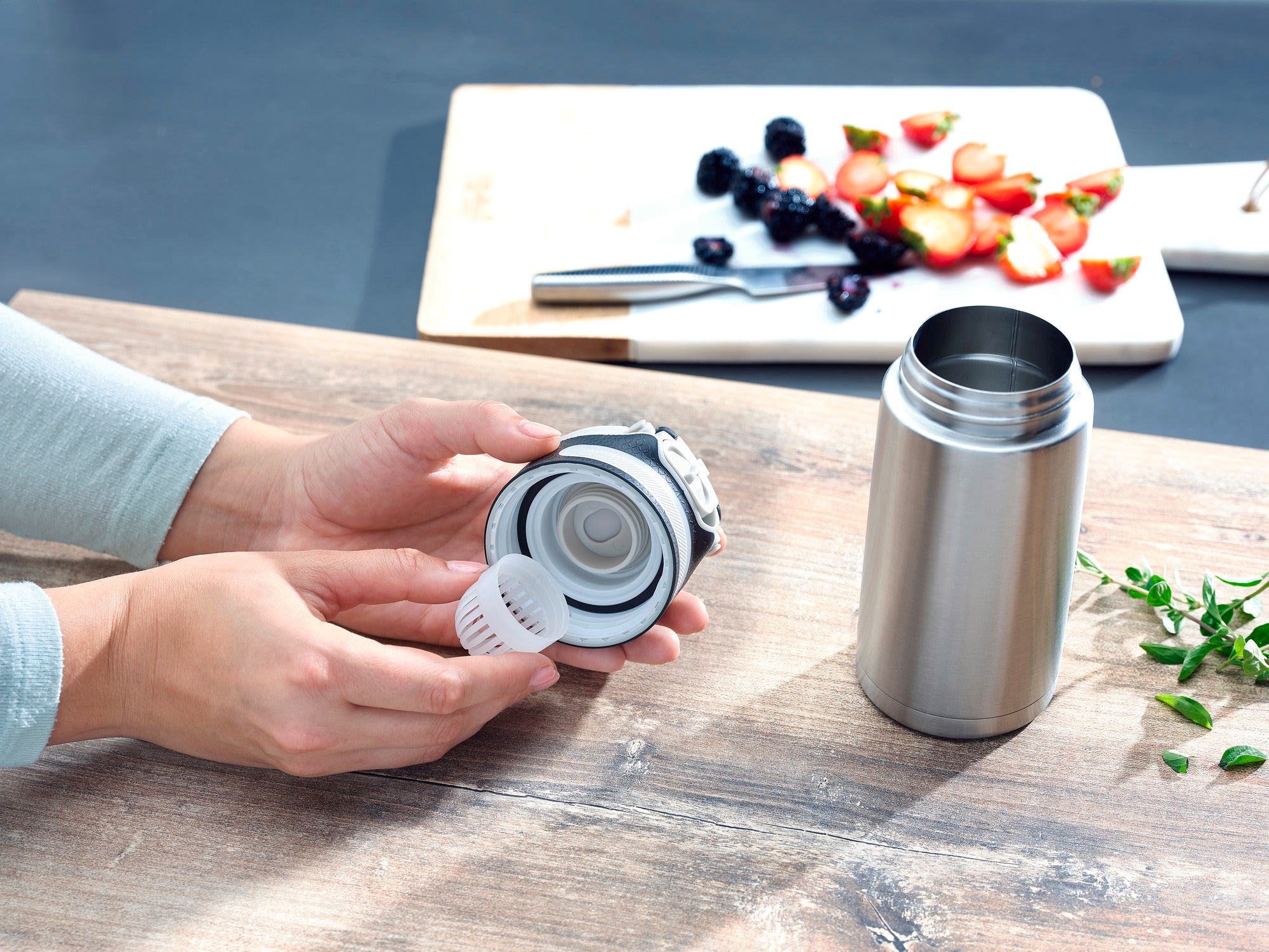 Le mug isotherme Flip 350 ml en argent est présenté sur une table en bois. Une personne tient le couvercle du mug, tandis qu'à l'arrière-plan, des fruits frais sont disposés sur une planche à découper.