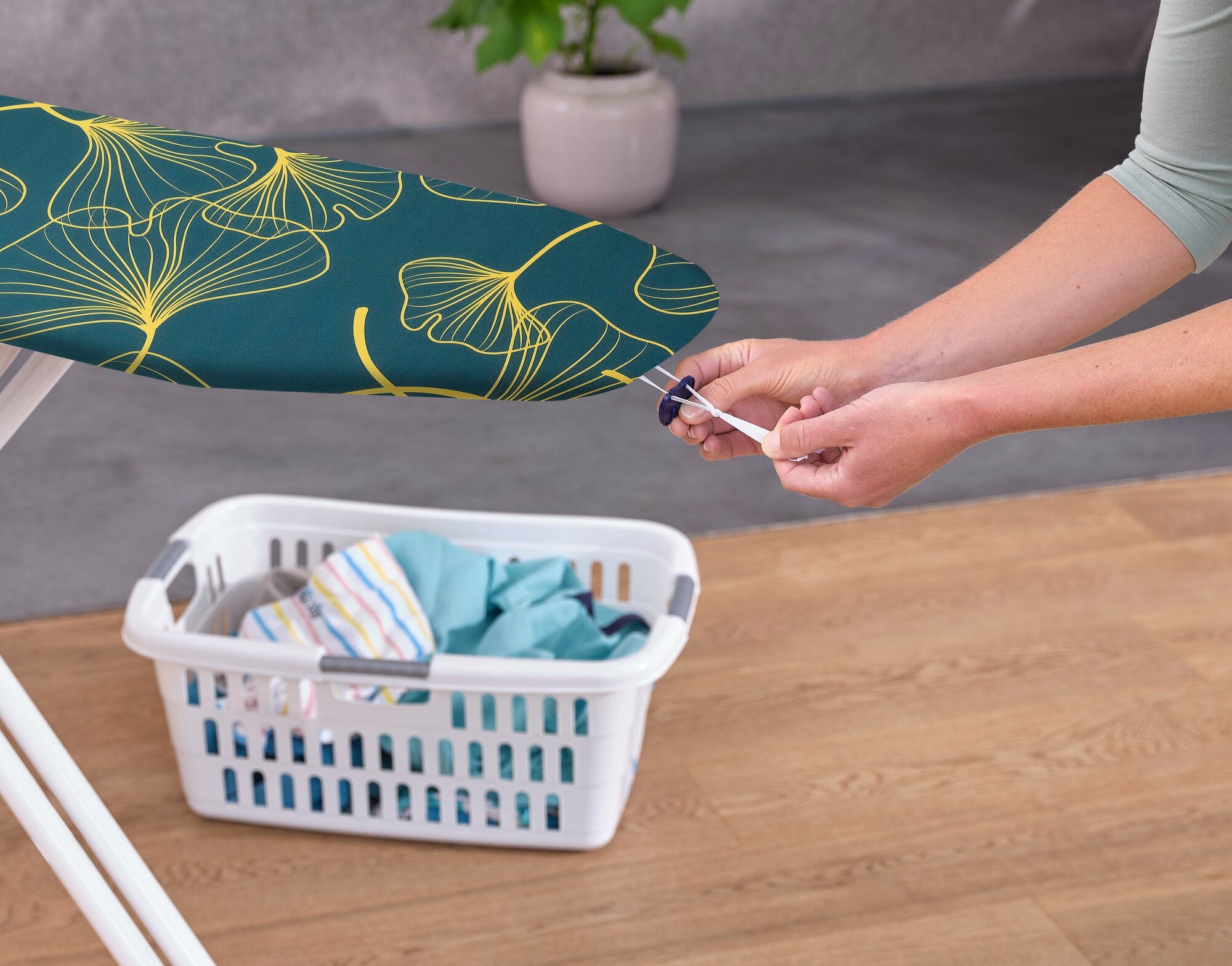 Eine Person zieht den Leifheit Bügeltischbezug Cotton Comfort S/M straff. Der Bezug hat ein modernes, grünes Design mit gelben Linien. Im Hintergrund steht ein Wäschekorb mit Kleidung.