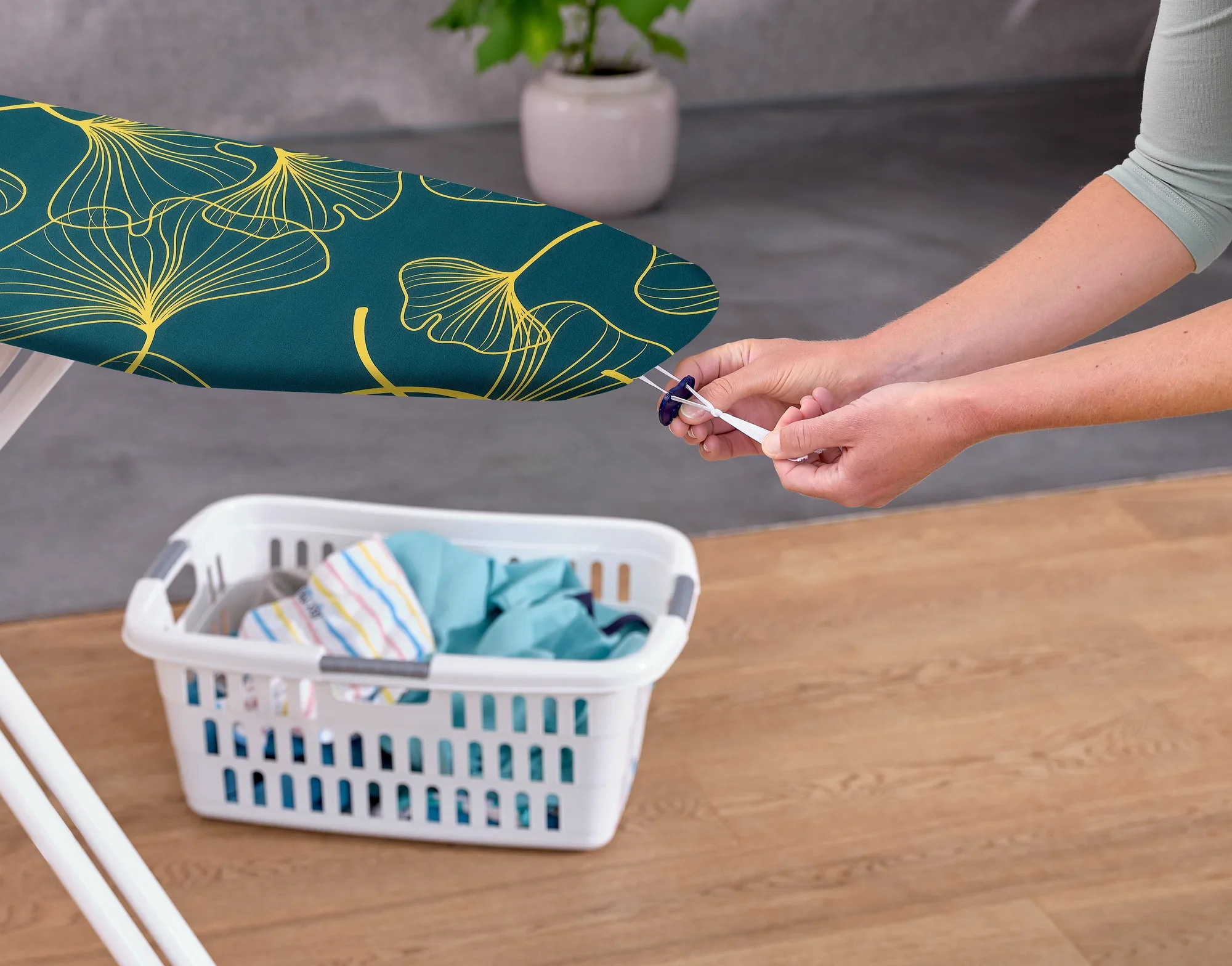 Eine Person zieht den Leifheit Bügeltischbezug Cotton Comfort S/M straff. Der Bezug hat ein modernes, grünes Design mit gelben Linien. Im Hintergrund steht ein Wäschekorb mit Kleidung.