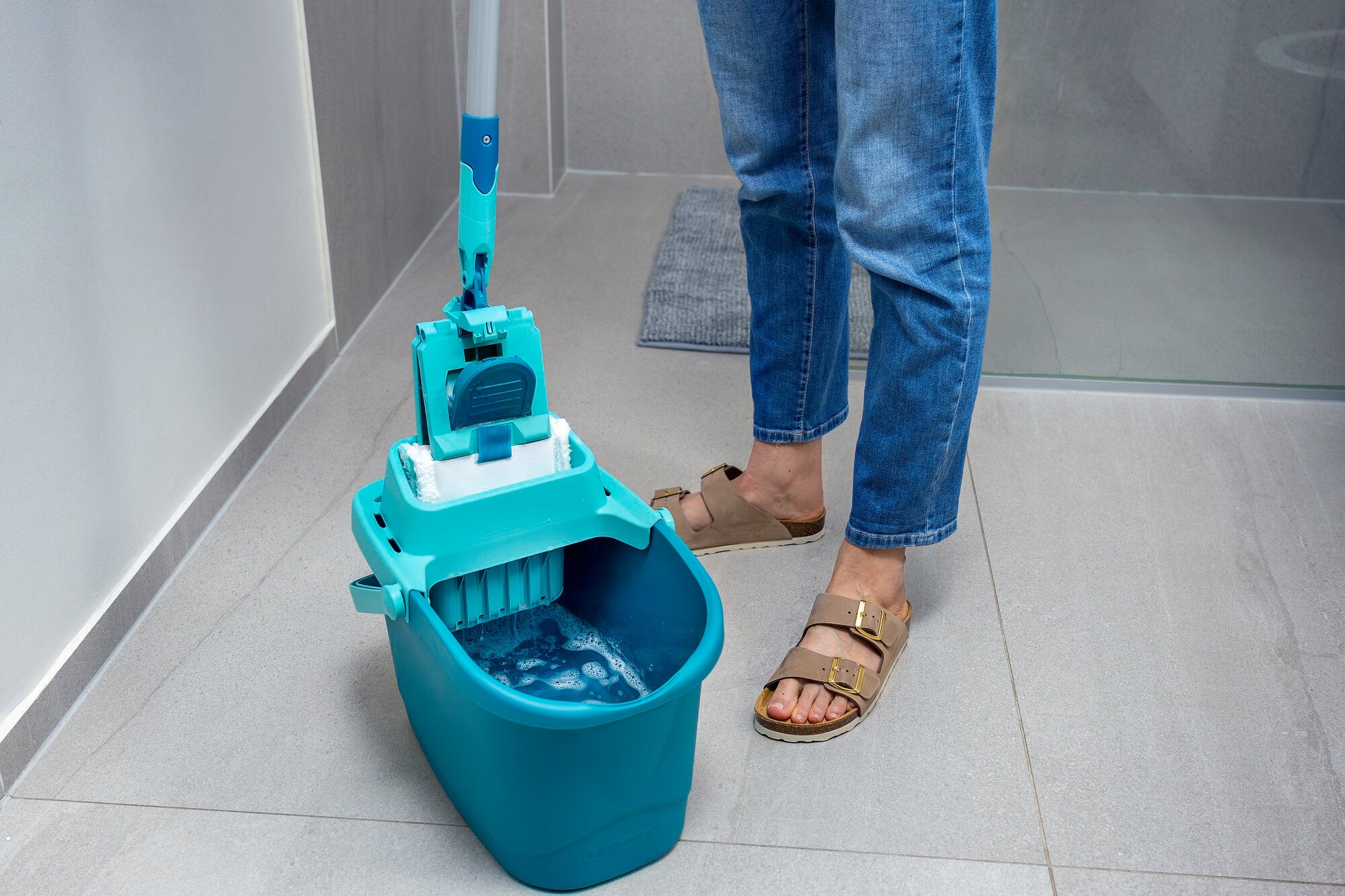 Una persona utiliza la mopa POWER CLEAN en un cuarto de baño moderno. El cubo está lleno de agua y la mopa está lista para limpiar. Ideal para una limpieza a fondo.