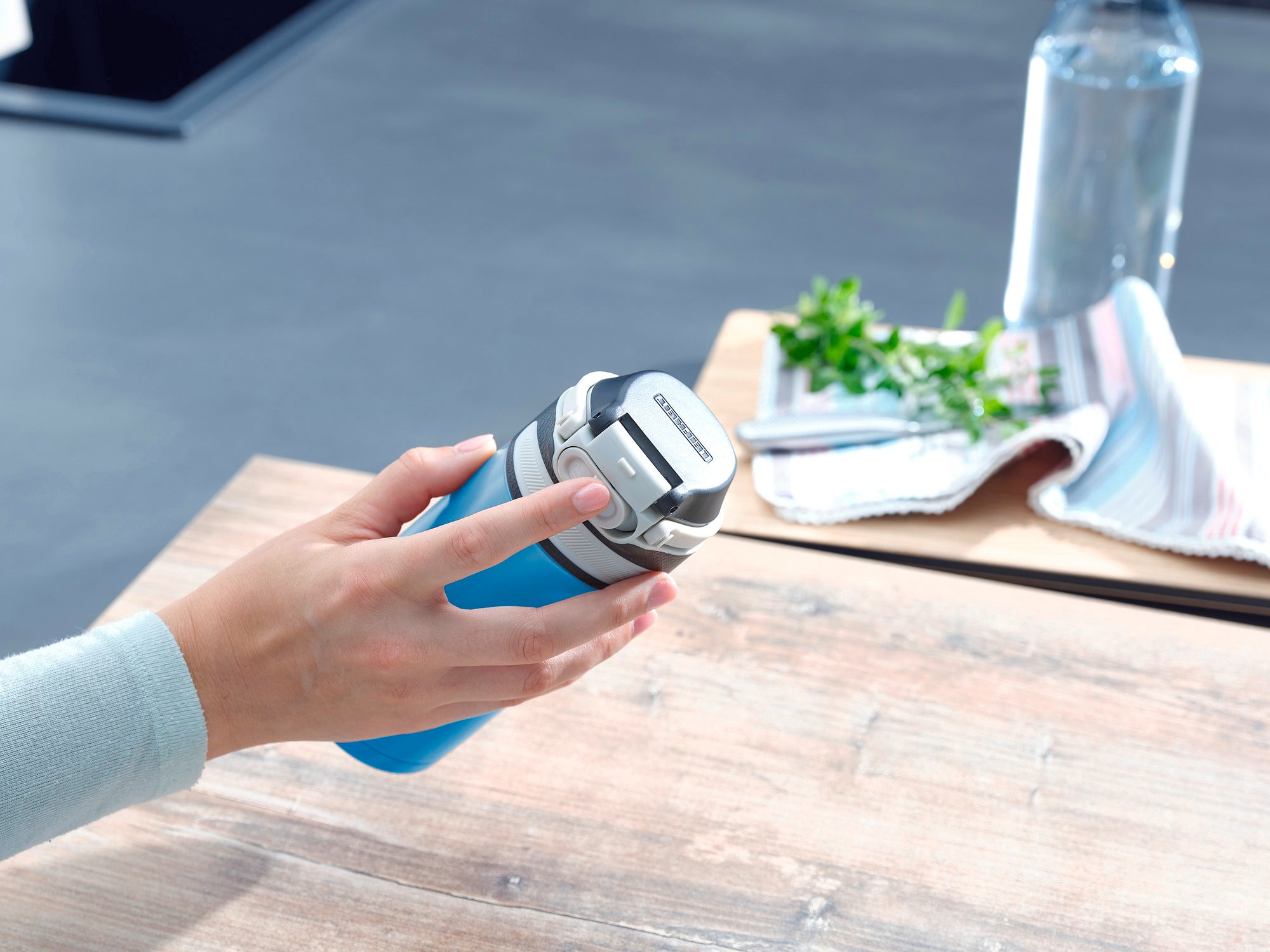 Eén persoon houdt een lichtblauwe Flip vacuümmok met een inhoud van 350 ml vast. De mok heeft een zilveren deksel en ligt op een houten tafel. Op de achtergrond zie je een handdoek en een fles water.