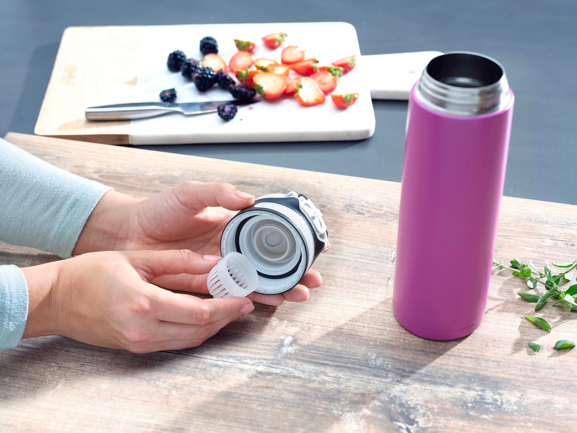 A purple Flip insulated mug with a capacity of 600 ml stands on a table. A person holds the lid of the mug, while sliced fruit can be seen in the background.