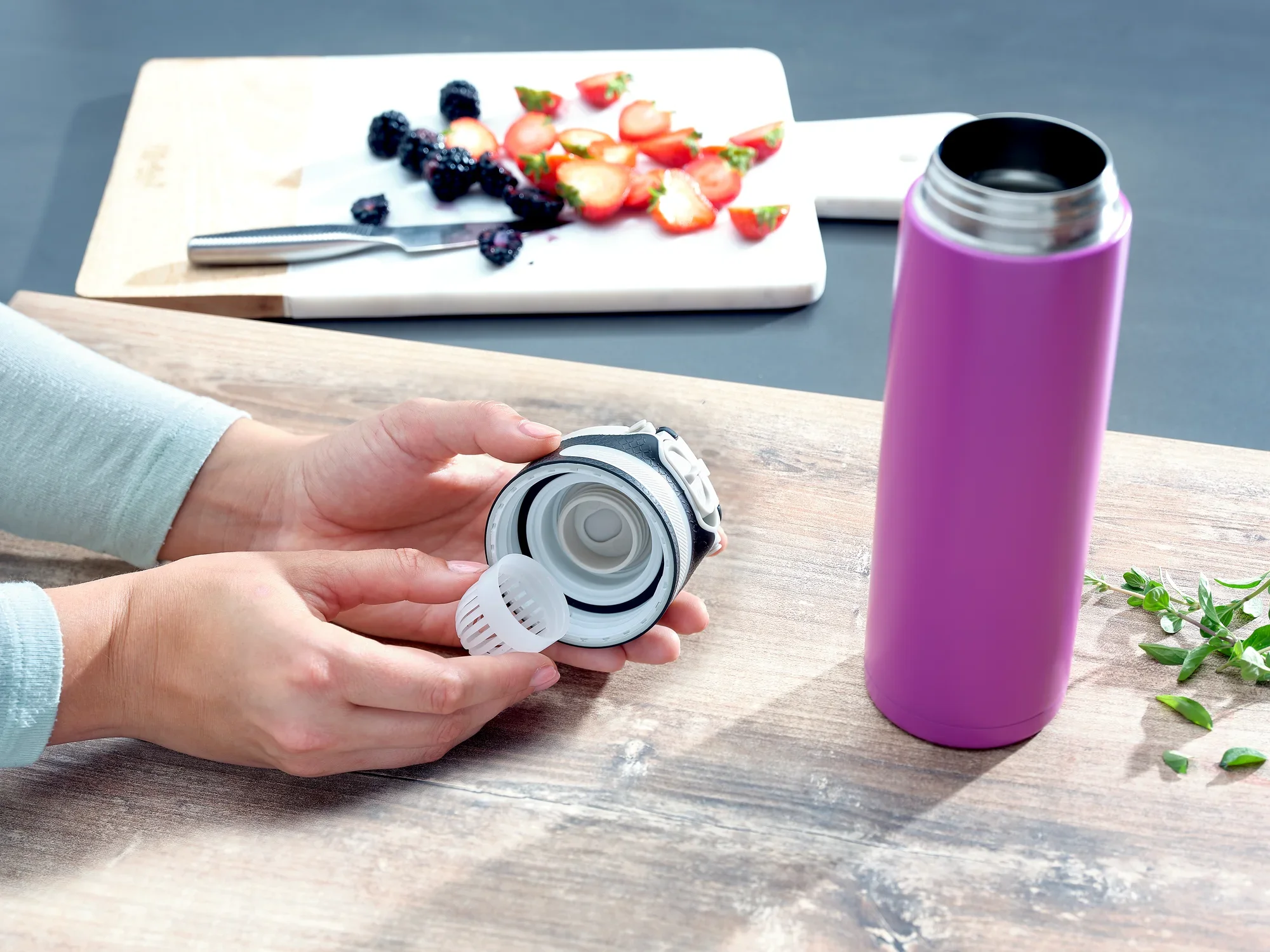 Een paarse Flip thermosfles met een inhoud van 600 ml staat op een tafel. Een persoon houdt het deksel van de mok vast, terwijl op de achtergrond gesneden fruit te zien is.