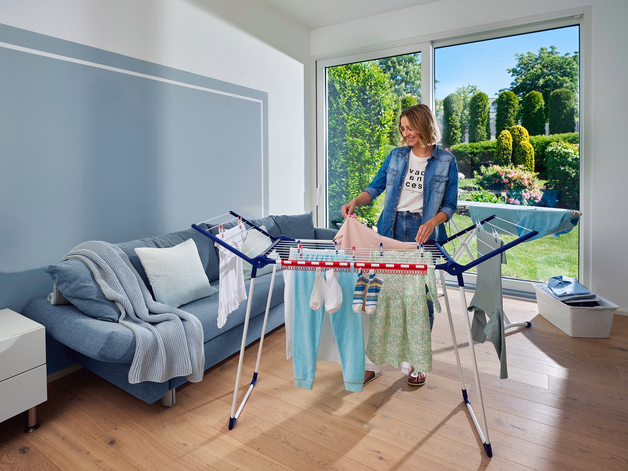 Een vrouw staat in een lichte woonkamer en hangt kleding op een Pegasus 150 Solid Slim wasdroger. Op de achtergrond is een goed onderhouden tuin te zien.