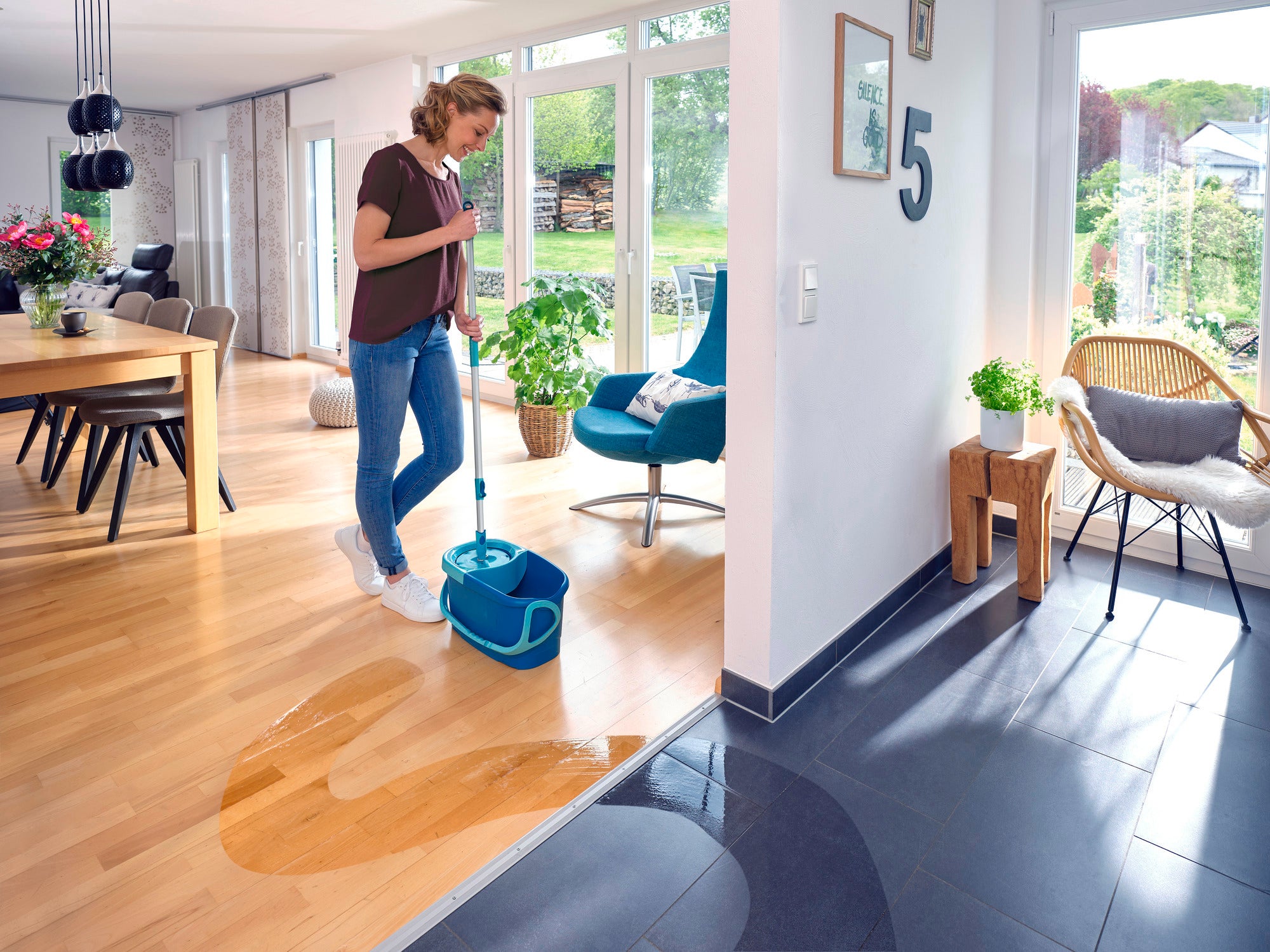 Une femme nettoie le parquet d'un salon moderne avec le système CLEAN TWIST. La pièce est lumineuse et élégamment meublée, avec de grandes fenêtres et une table à manger à l'arrière-plan.