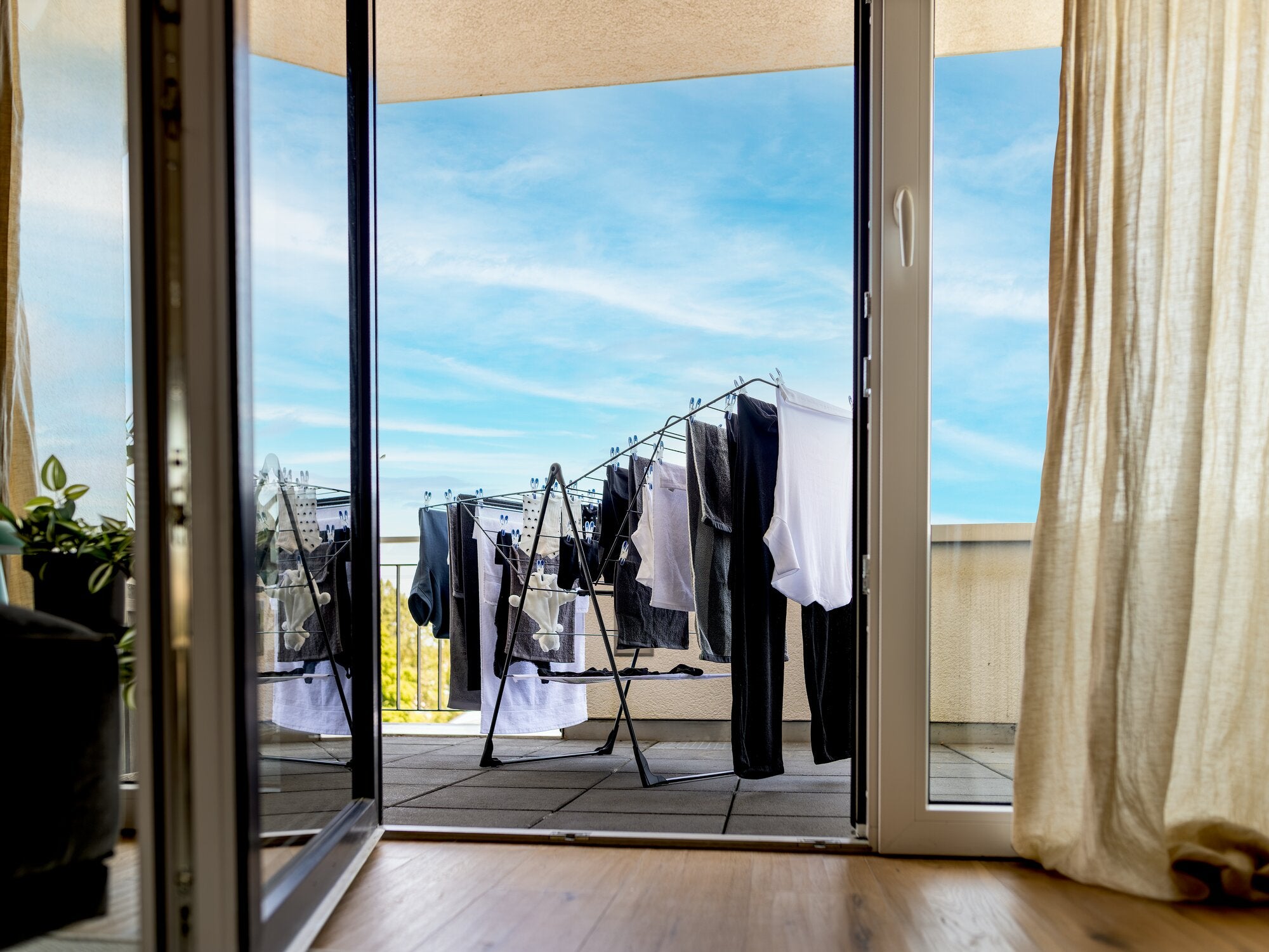 Leifheit Wäscheschirm Classic 250 Flex Black auf einem Balkon vor blauem Himmel, bespannt mit dunkler und heller Wäsche; Blick durch eine geöffnete Balkontür in heller, wohnlicher Szene.