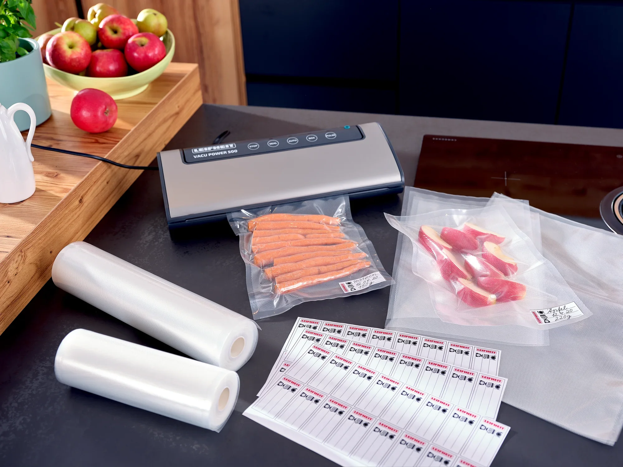Worek próżniowy 20x30 cm na stole kuchennym ze zgrzewarką próżniową, dwiema rolkami folii próżniowej, zapakowaną próżniowo żywnością i etykietami. Idealny do zachowania świeżości.
