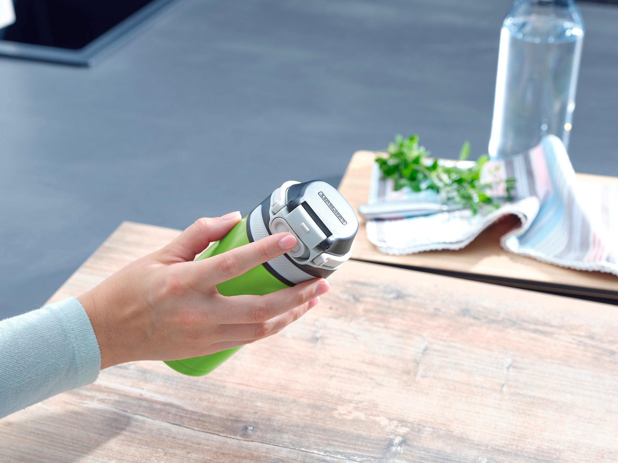 Eén persoon houdt een groene vacuümklapbeker met een inhoud van 350 ml vast. De mok heeft een zilveren deksel. Op de achtergrond zijn een theedoek en een waterfles te zien.