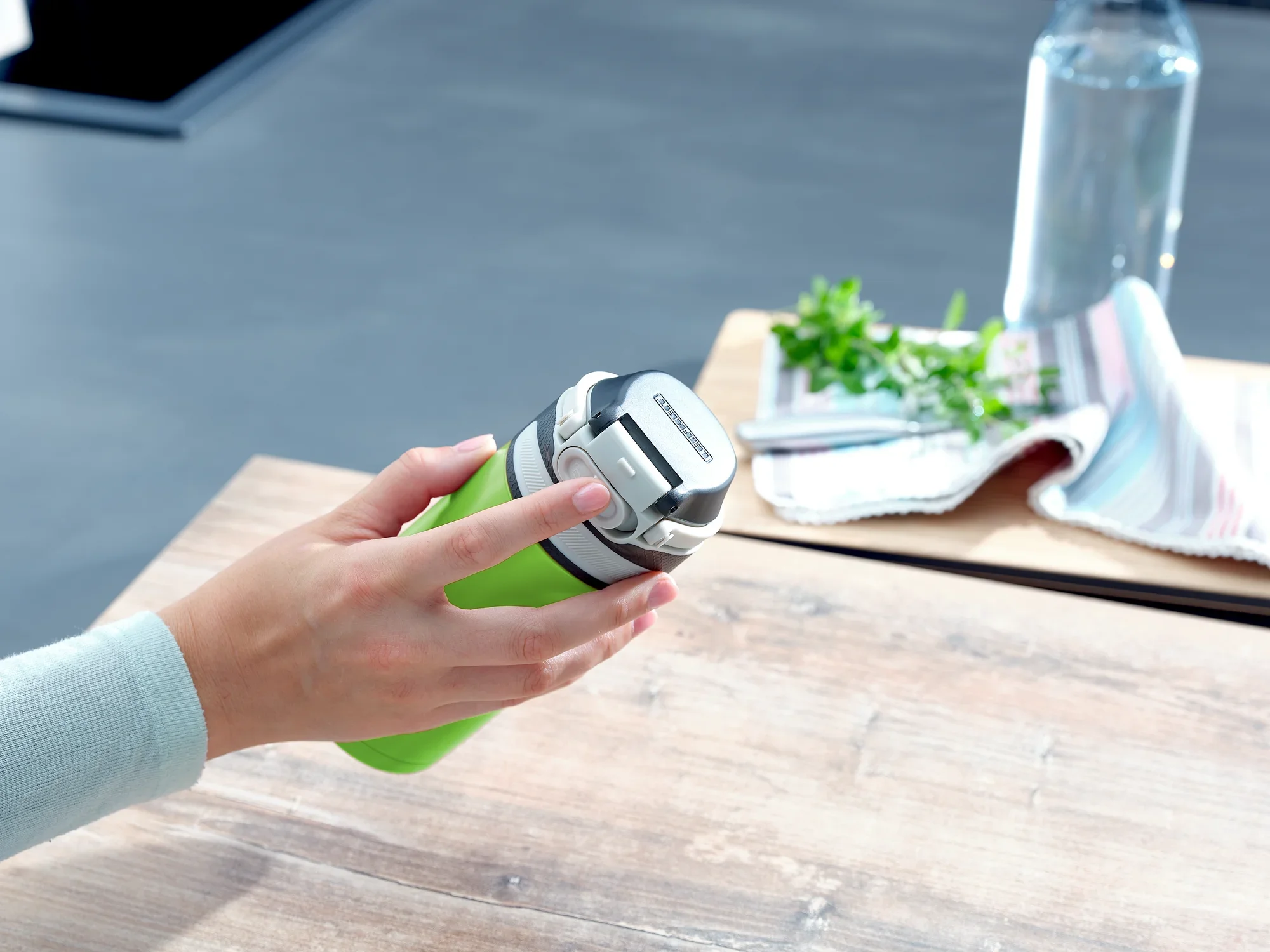 Jedna osoba trzyma zielony kubek próżniowy o pojemności 350 ml. Kubek ma srebrną pokrywkę. W tle widać ściereczkę i butelkę wody.