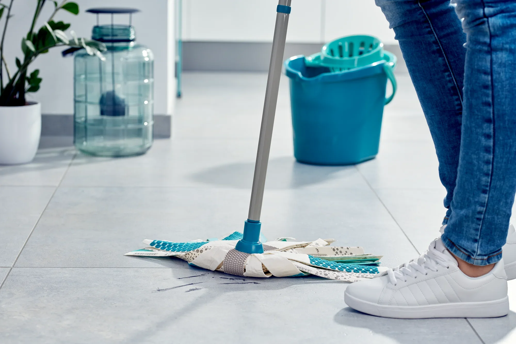 A person is cleaning a light-coloured tiled floor with the Power Mop 3in1. A blue bucket is standing in the background. The mop has a blue handle and white mopping strips.