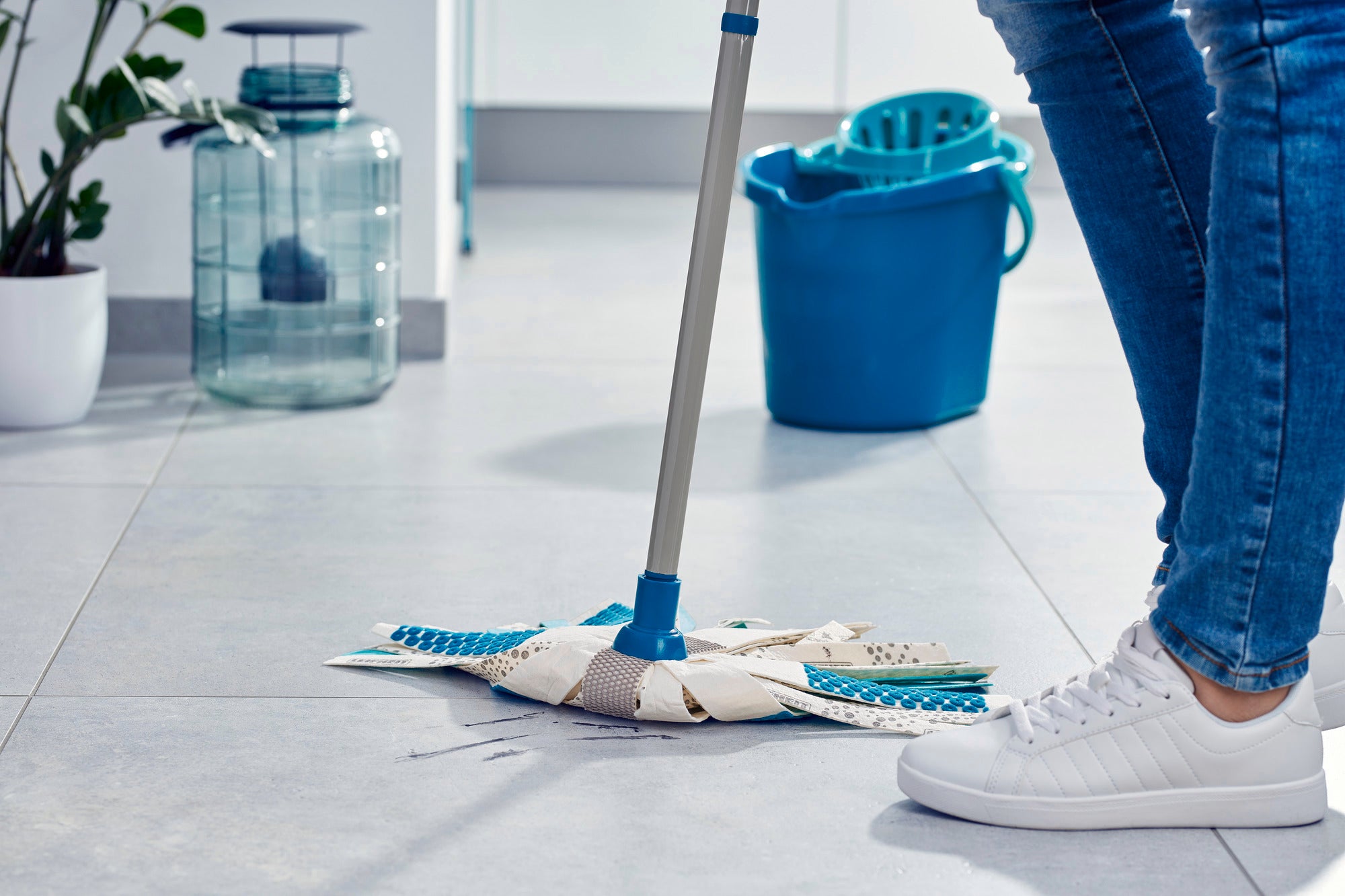 Une personne nettoie un sol carrelé clair avec le Power Mop 3in1. Un seau bleu se trouve à l'arrière-plan. La serpillière a un manche bleu et des bandes blanches.
