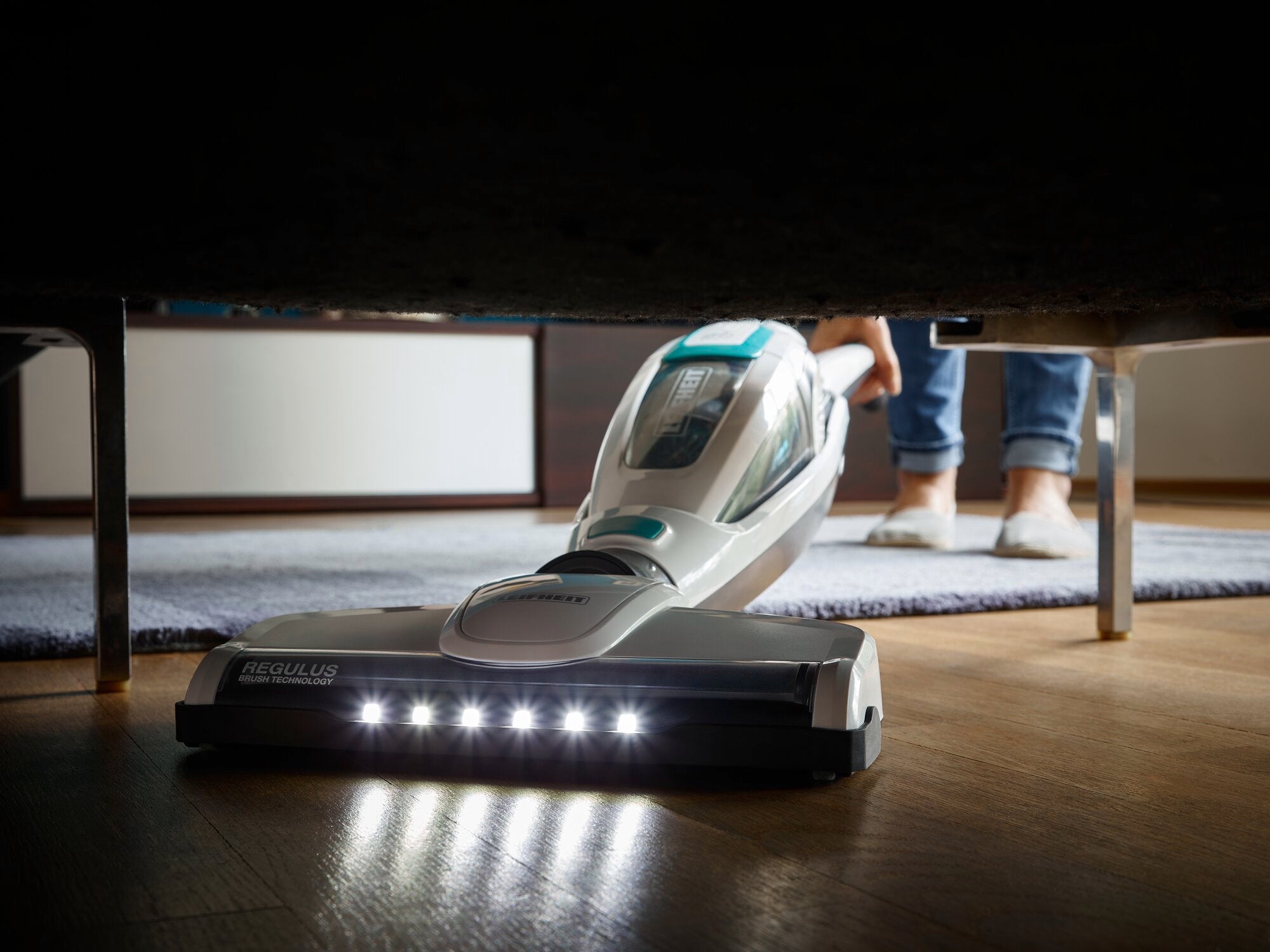 Una aspiradora Regulus PowerVac 2 en 1 limpia el suelo debajo de un sofá. Las luces LED de la parte delantera del aparato iluminan la zona, mientras una persona se encuentra de pie al fondo.