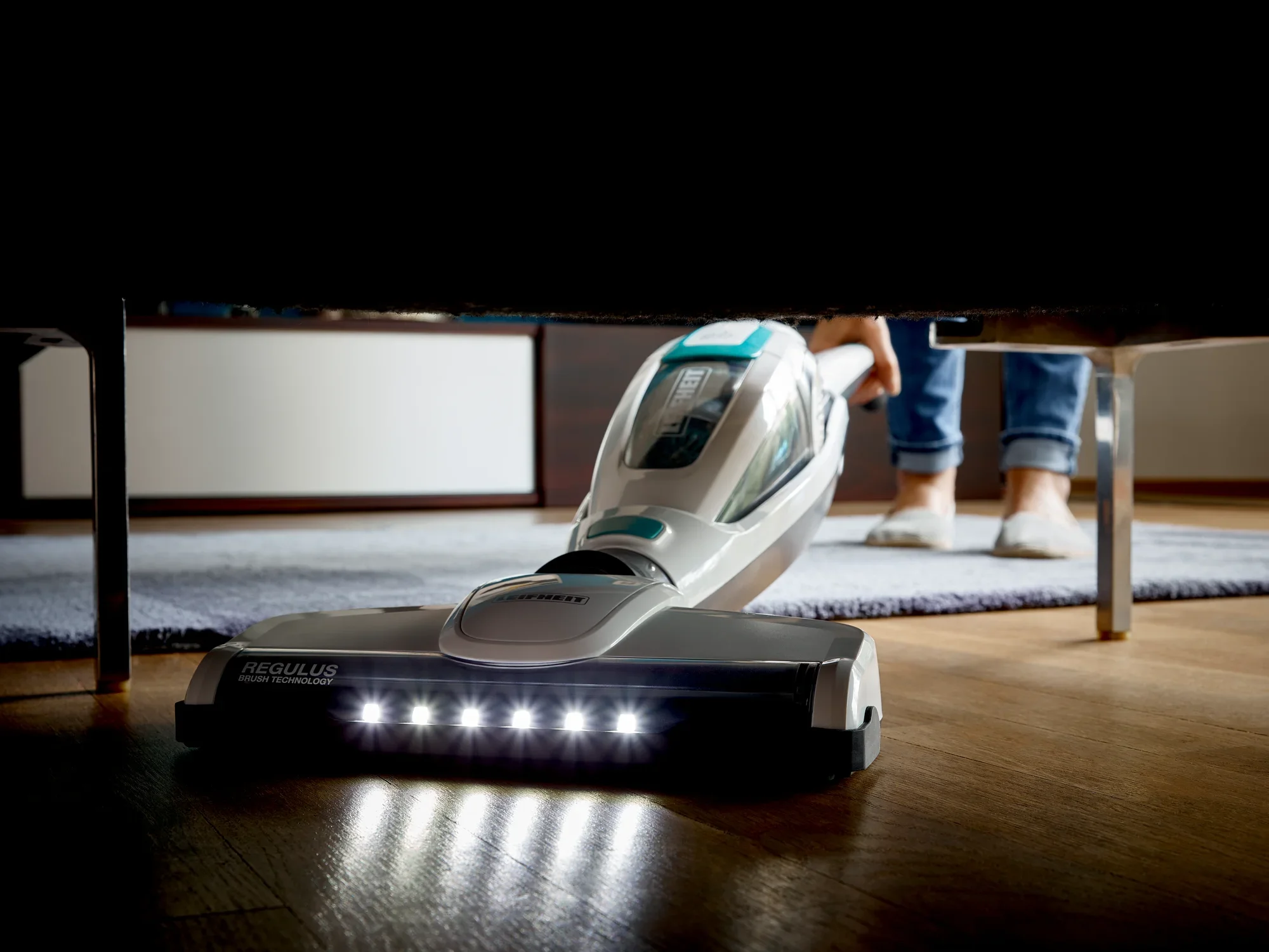 Un aspirateur Regulus PowerVac 2 en 1 nettoie le sol sous un canapé. Les LED à l'avant de l'appareil éclairent la zone tandis qu'une personne se tient à l'arrière.
