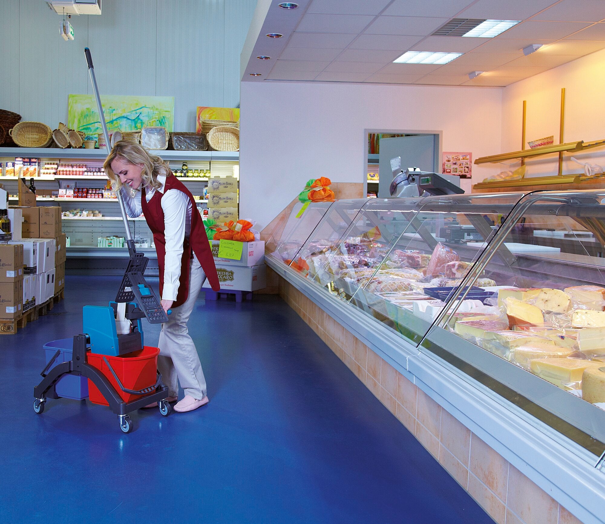 Une personne nettoie le sol d'un supermarché avec le système de nettoyage à plat Professional. À l'arrière-plan, on aperçoit des comptoirs réfrigérés avec divers produits alimentaires.