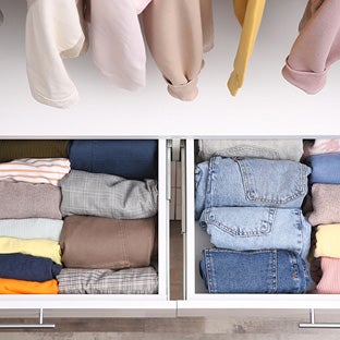 In een kast zijn kledinghangers met beige en gele kledingstukken boven lades met netjes opgevouwen truien en jeans zichtbaar.