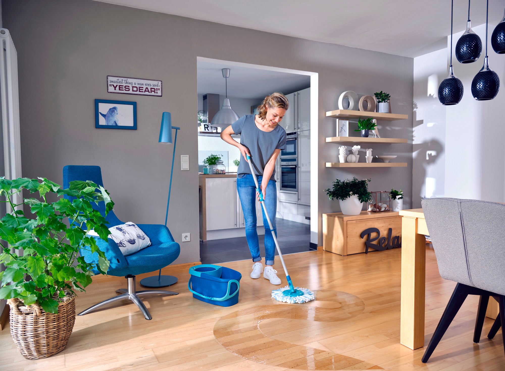 Kobieta czyści drewnianą podłogę w nowoczesnym salonie za pomocą CLEAN TWIST Mop Ergo. Pokój jest jasny i stylowo urządzony, z roślinami i nowoczesnymi meblami.
