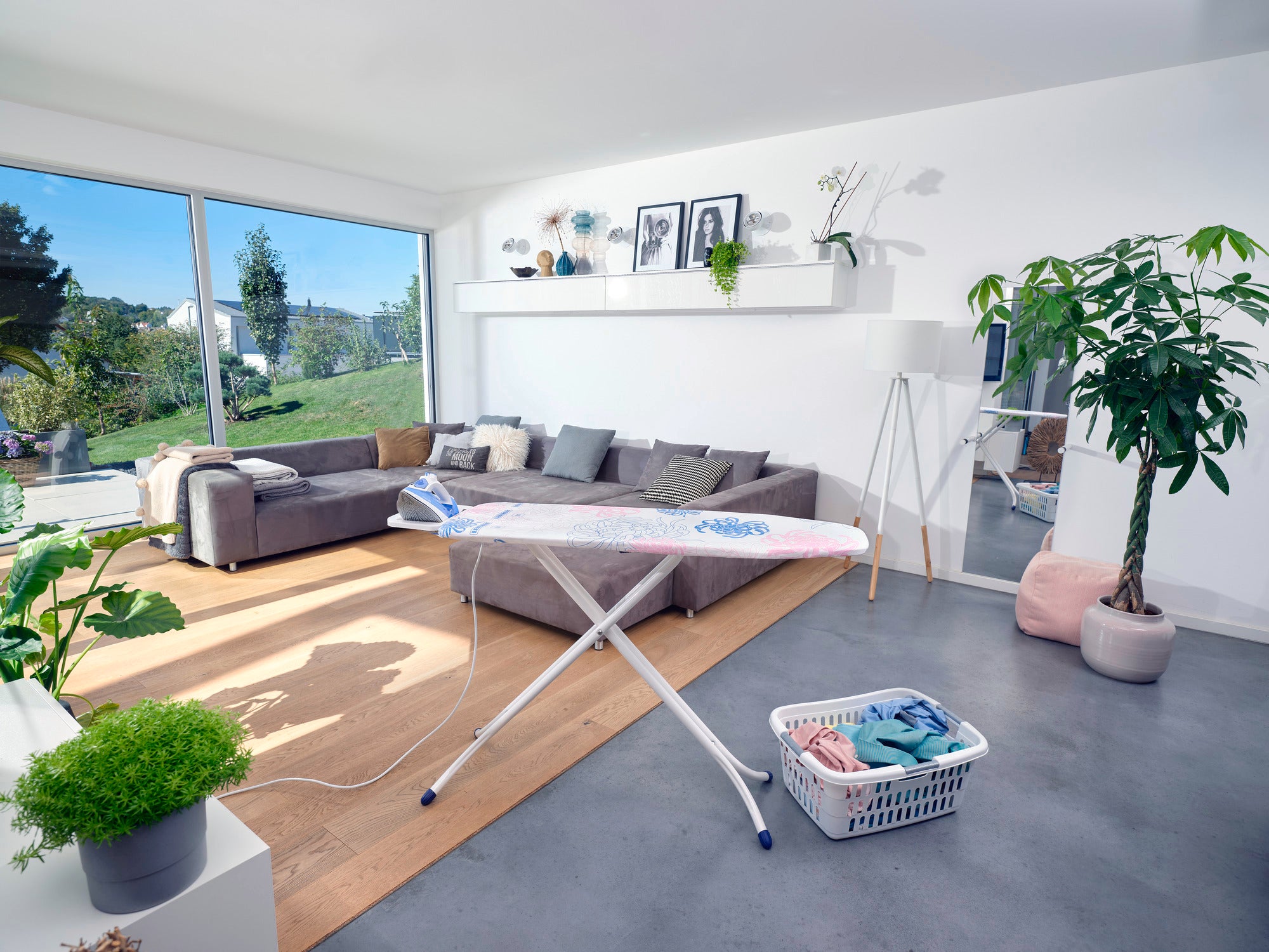 Bügeltisch Classic M Compact in einem modernen Wohnzimmer mit großen Fenstern. Der Tisch steht auf einem grauen Boden, umgeben von Pflanzen und einem Korb mit Wäsche.