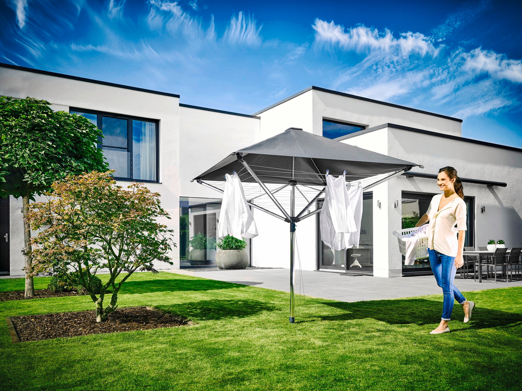 Een vrouw staat in een tuin naast de LinoProtect 400 droogmolen, die open staat en vol hangt met wit wasgoed. Op de achtergrond is een modern huis te zien.