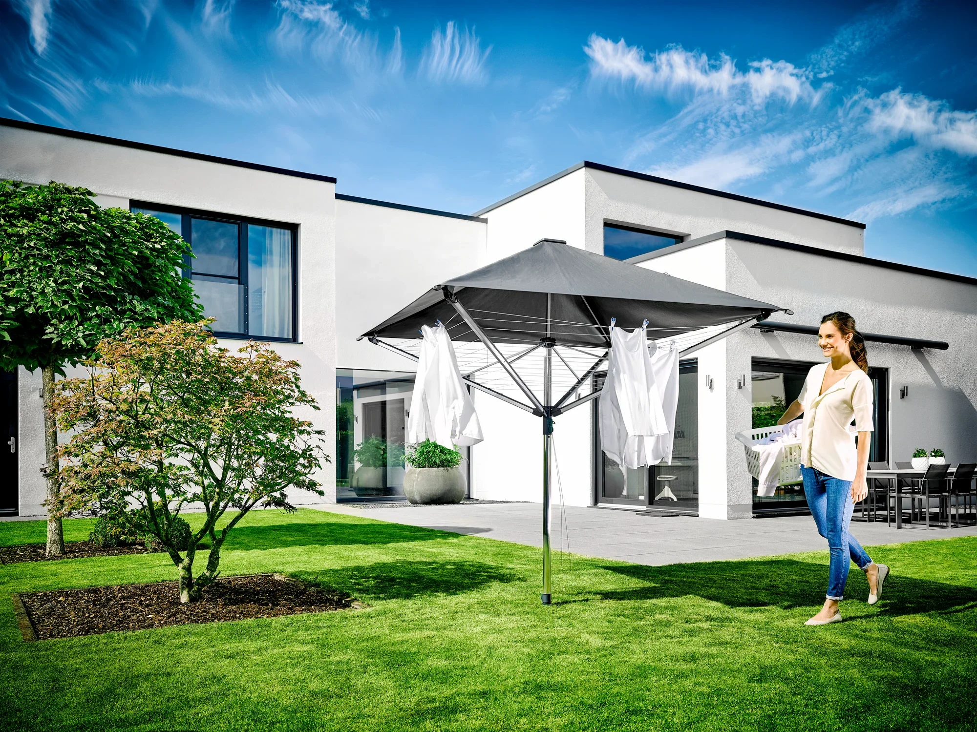 Une femme se tient dans un jardin à côté du séchoir à linge LinoProtect 400. Le séchoir est ouvert et recouvert de linge blanc. Une maison moderne est visible en arrière-plan.