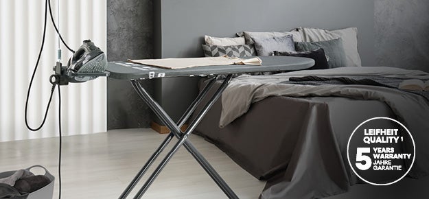 Buegeleisen und Buegelbrett stehen in einem modern eingerichteten Schlafzimmer mit grauen Bettdecken und Dekokissen, 5-Jahre-Garantie-Logo.