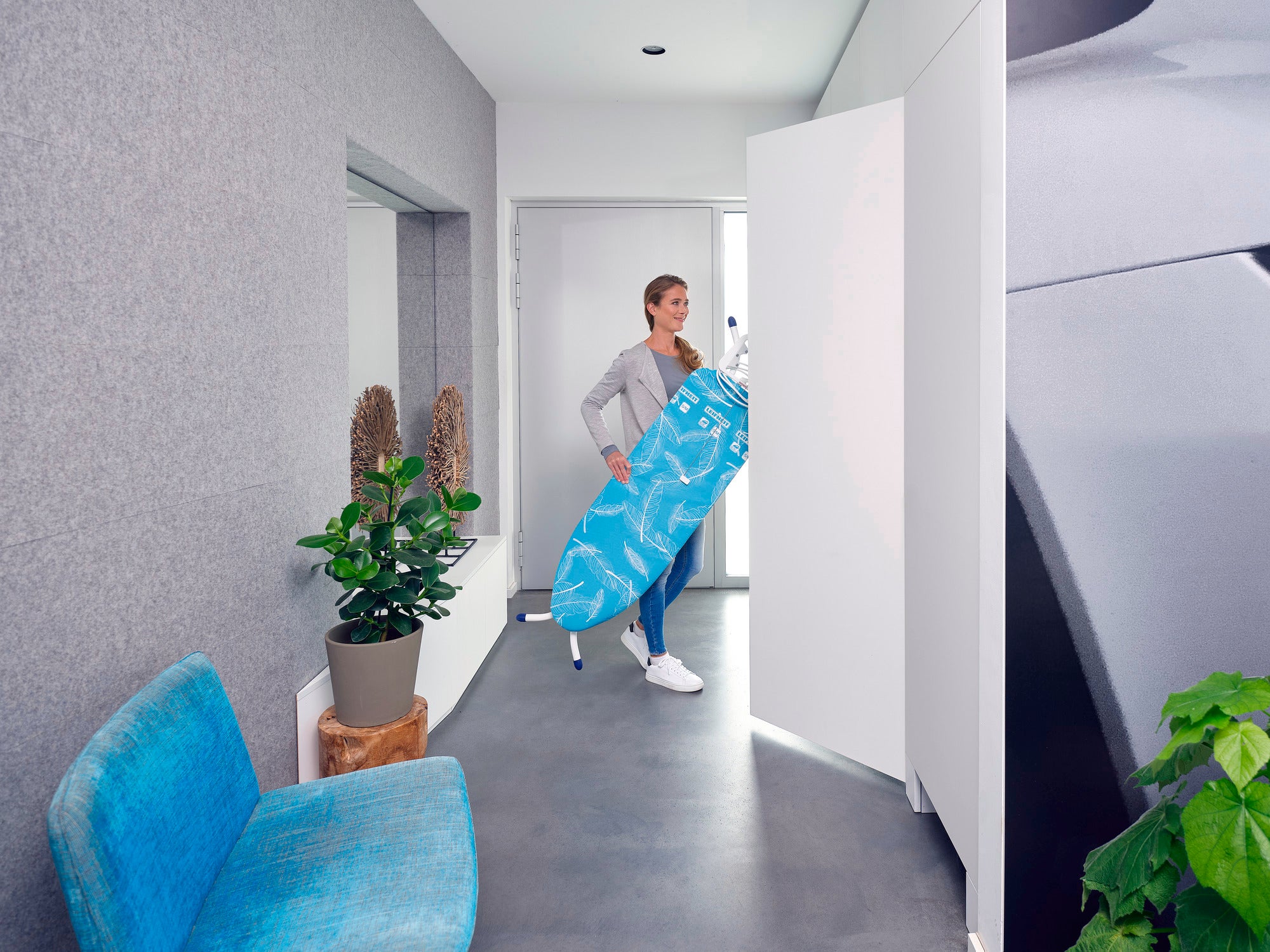 Eine Frau trägt den Bügeltisch Air Board M Compact Plus durch einen modernen Flur. Der Bügeltisch ist leicht und handlich, ideal für den einfachen Transport und die platzsparende Aufbewahrung.