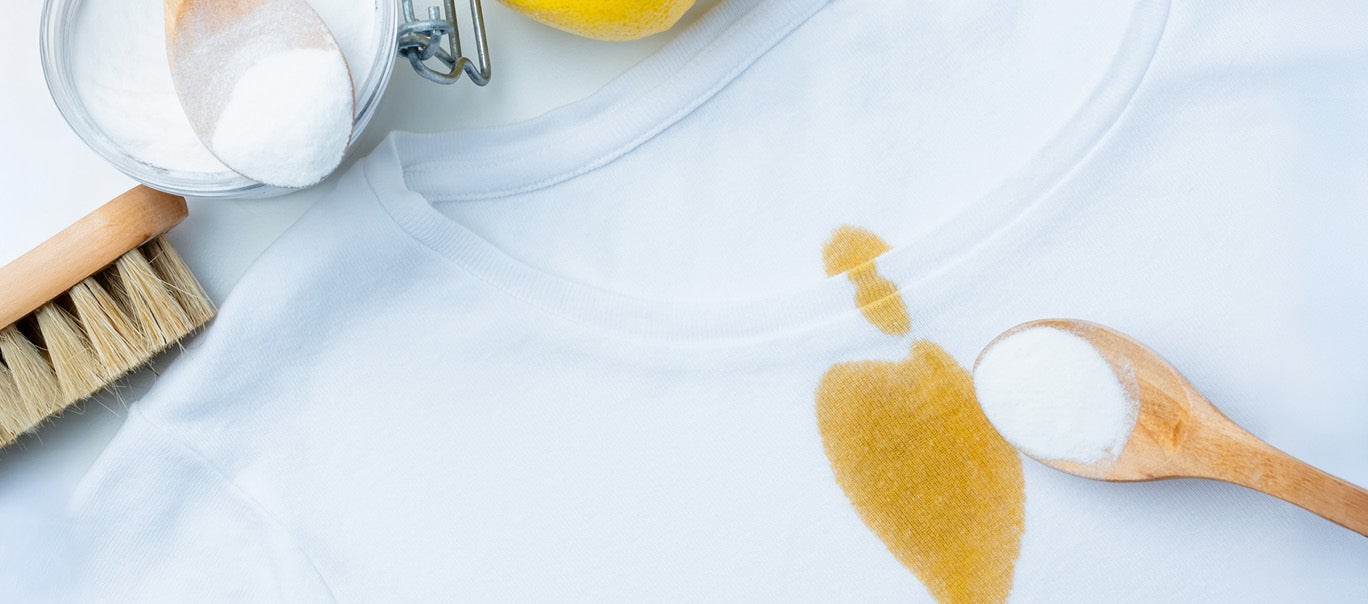 Flecken entfernen – so wird Kleidung im Nu sauber Weisses T-Shirt mit gelbem Fleck, daneben Buerste, Zitrone, Holzloeffel mit Pulver und Schale, die Reinigung andeuten.