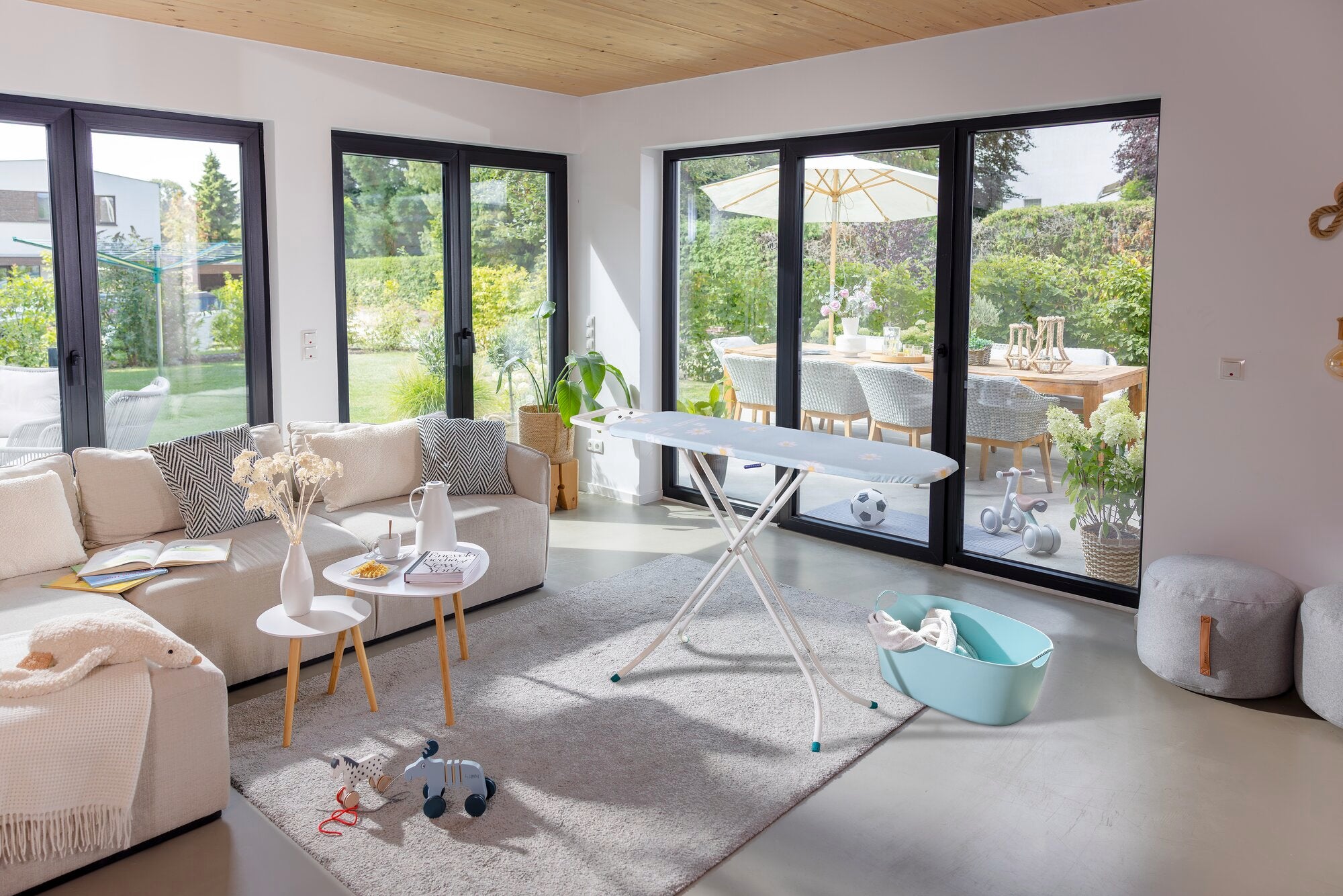 Bügelbrett Leifheit Classic S Basic in einem modernen Wohnzimmer mit großen Fenstern. Das Brett steht auf einem Teppich, umgeben von einem Sofa und einem Tisch mit Dekoration.