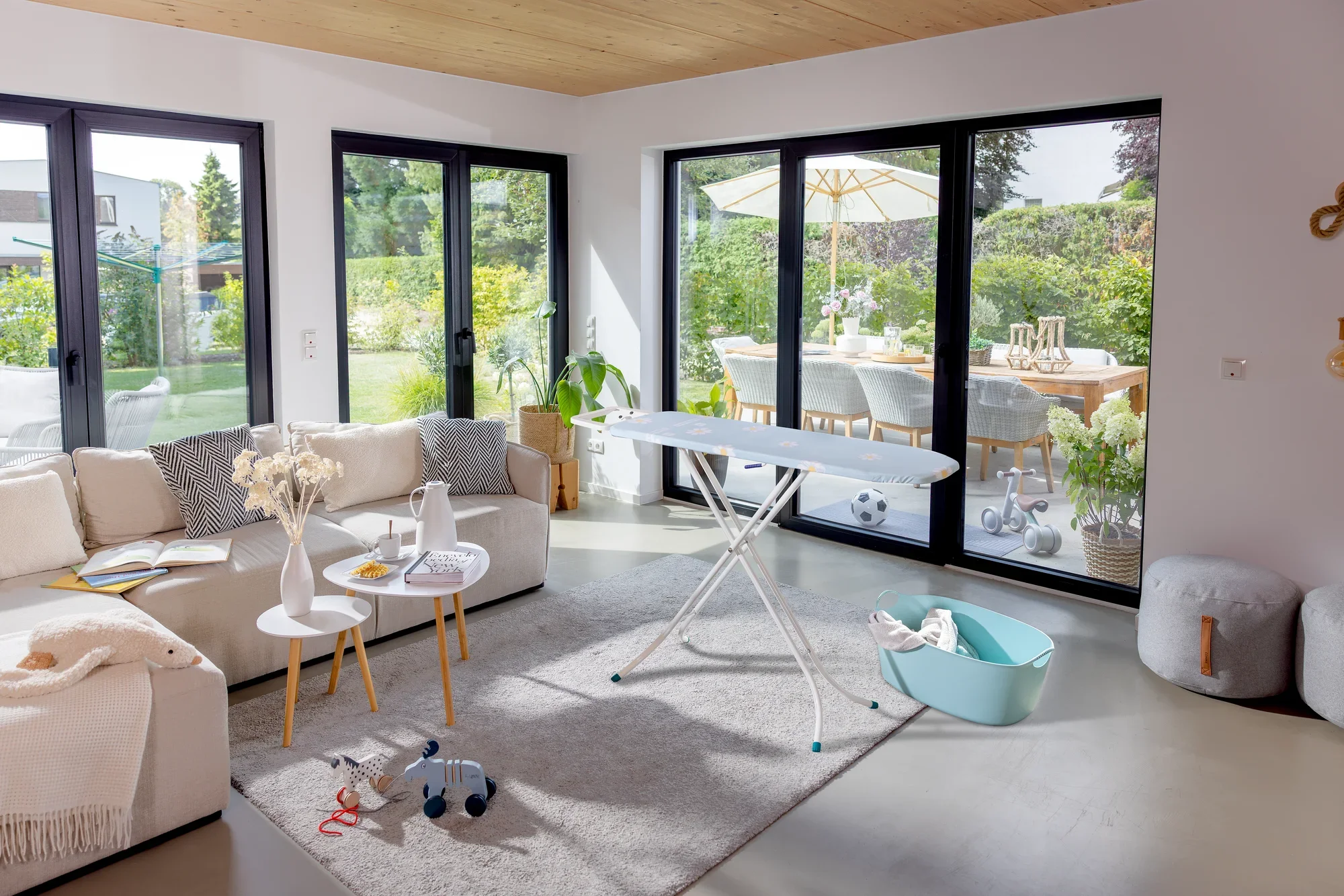 Bügelbrett Leifheit Classic S Basic in einem modernen Wohnzimmer mit großen Fenstern. Das Brett steht auf einem Teppich, umgeben von einem Sofa und einem Tisch mit Dekoration.