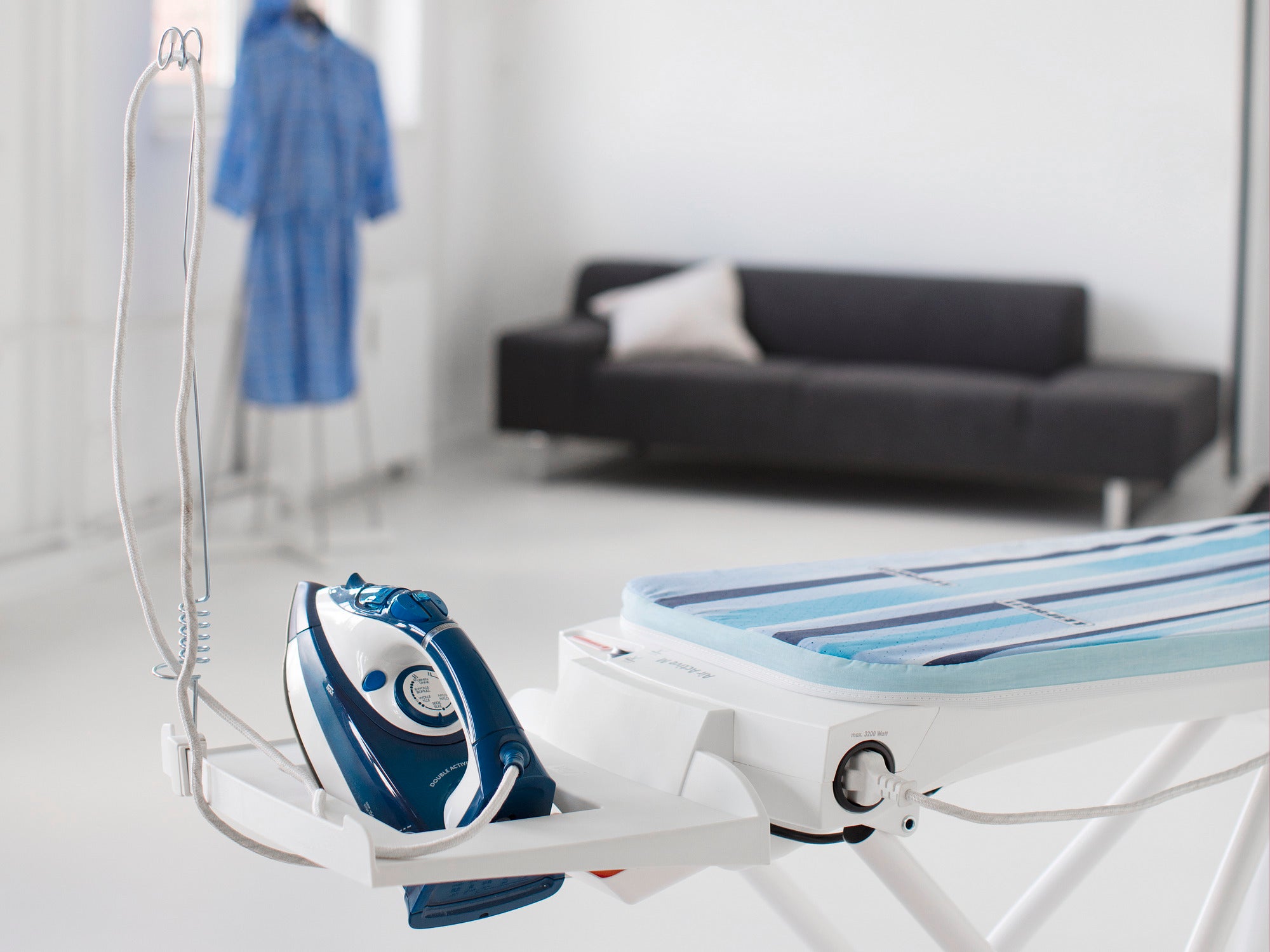 Air Active Express M ironing table in white with integrated steam iron. In the background, a sofa and a blue bathrobe on a stand. Ideal for efficient ironing.