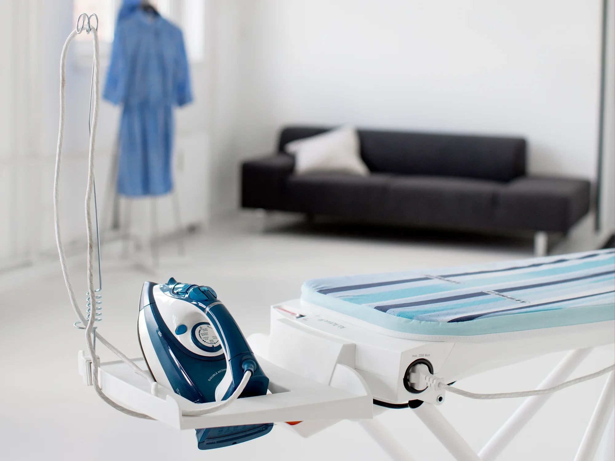 Table à repasser Air Active Express M en blanc avec fer à vapeur intégré. À l'arrière-plan, un canapé et un peignoir bleu sur un porte-serviette. Idéale pour un repassage efficace.