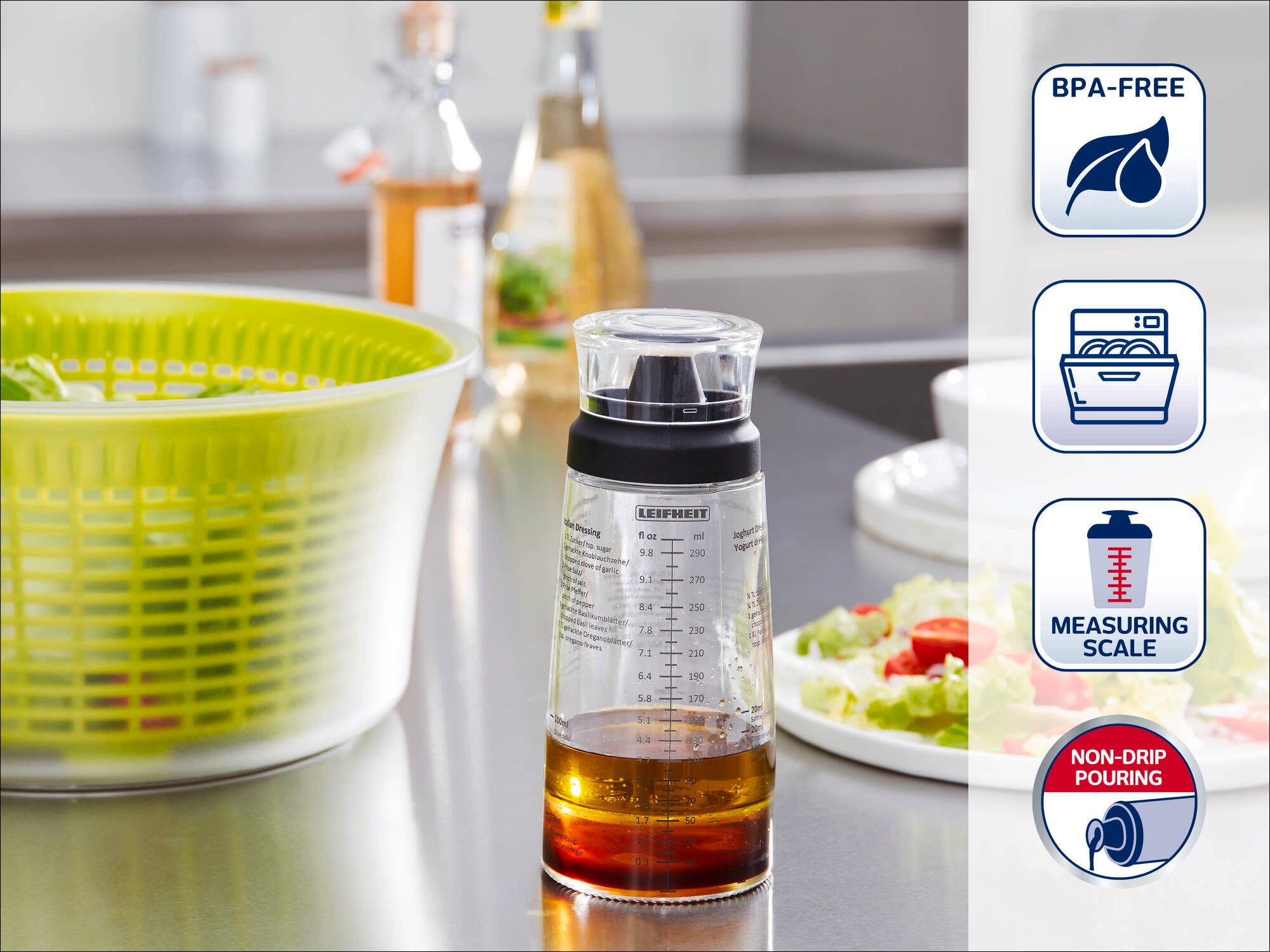 Ein transparenter Messbecher mit schwarzem Deckel steht auf einer Küchenarbeitsplatte. Er zeigt eine Messskala und ist BPA-frei. Im Hintergrund sind eine Salatschleuder und frische Zutaten zu sehen.