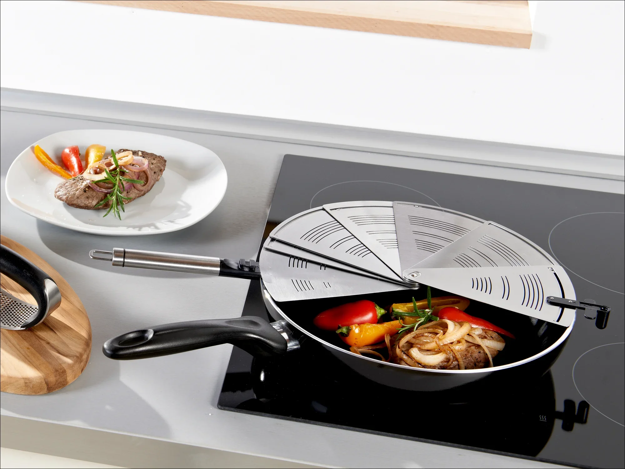 Une poêle avec un couvercle anti-éclaboussures sur une cuisinière. La poêle contient des poivrons colorés et des oignons. À l'arrière-plan, on aperçoit une assiette avec un steak.