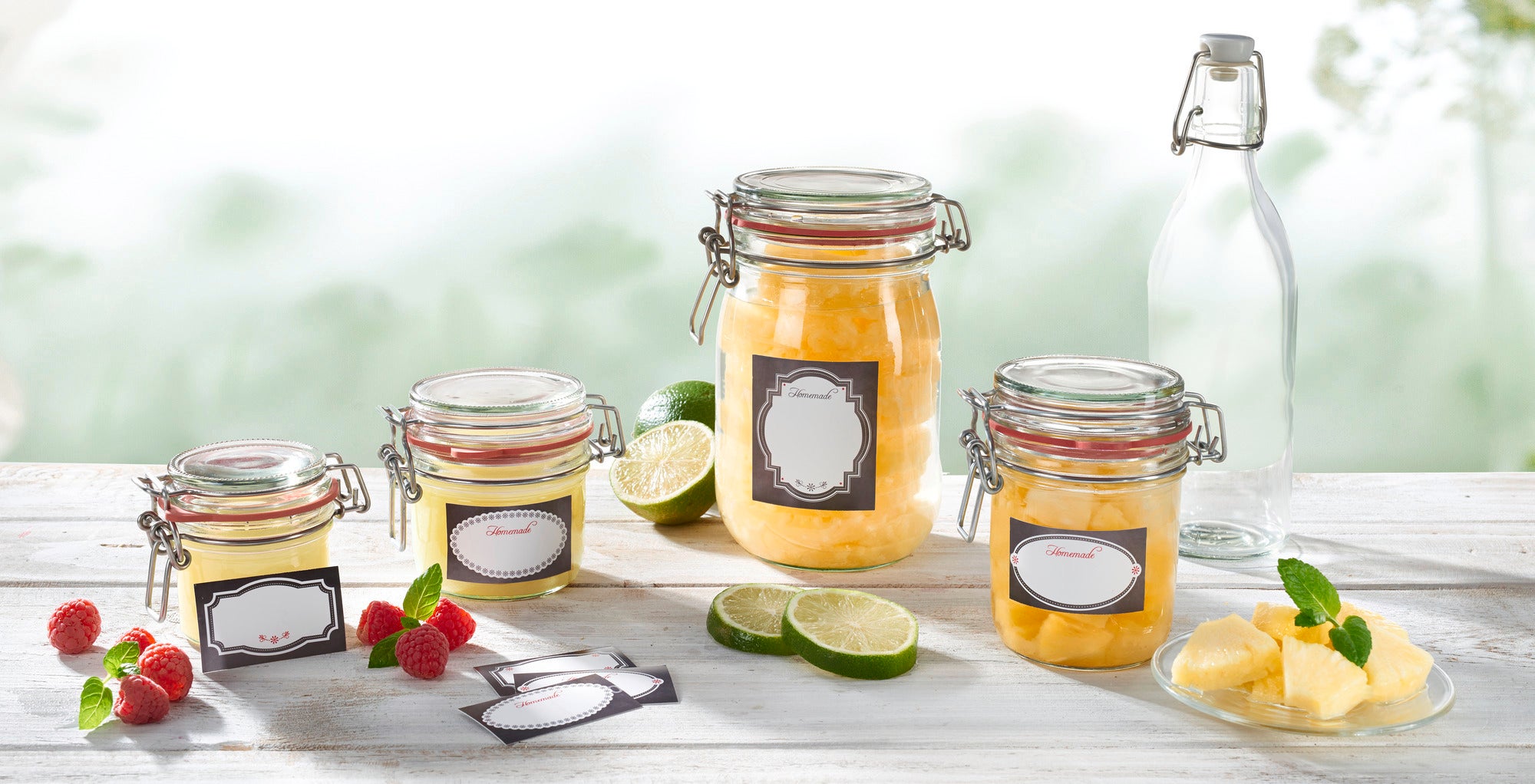 Vier Einmachgläser mit dekorativen schwarz-weißen Etiketten stehen auf einem Tisch. Sie sind mit gelbem Inhalt gefüllt, umgeben von Limetten, Himbeeren und Ananasstücken.